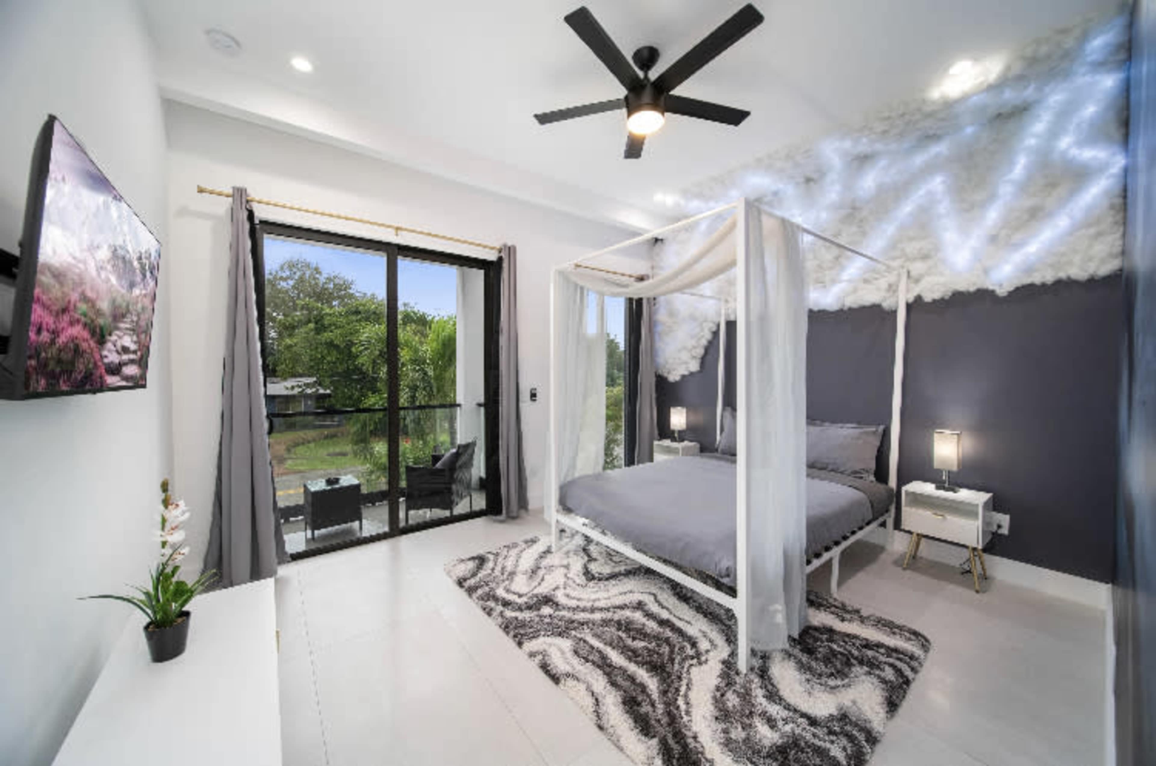 The image shows a modern bedroom with a four-poster bed, a ceiling fan, a television mounted on the wall, and large windows that open to a view of greenery outside.