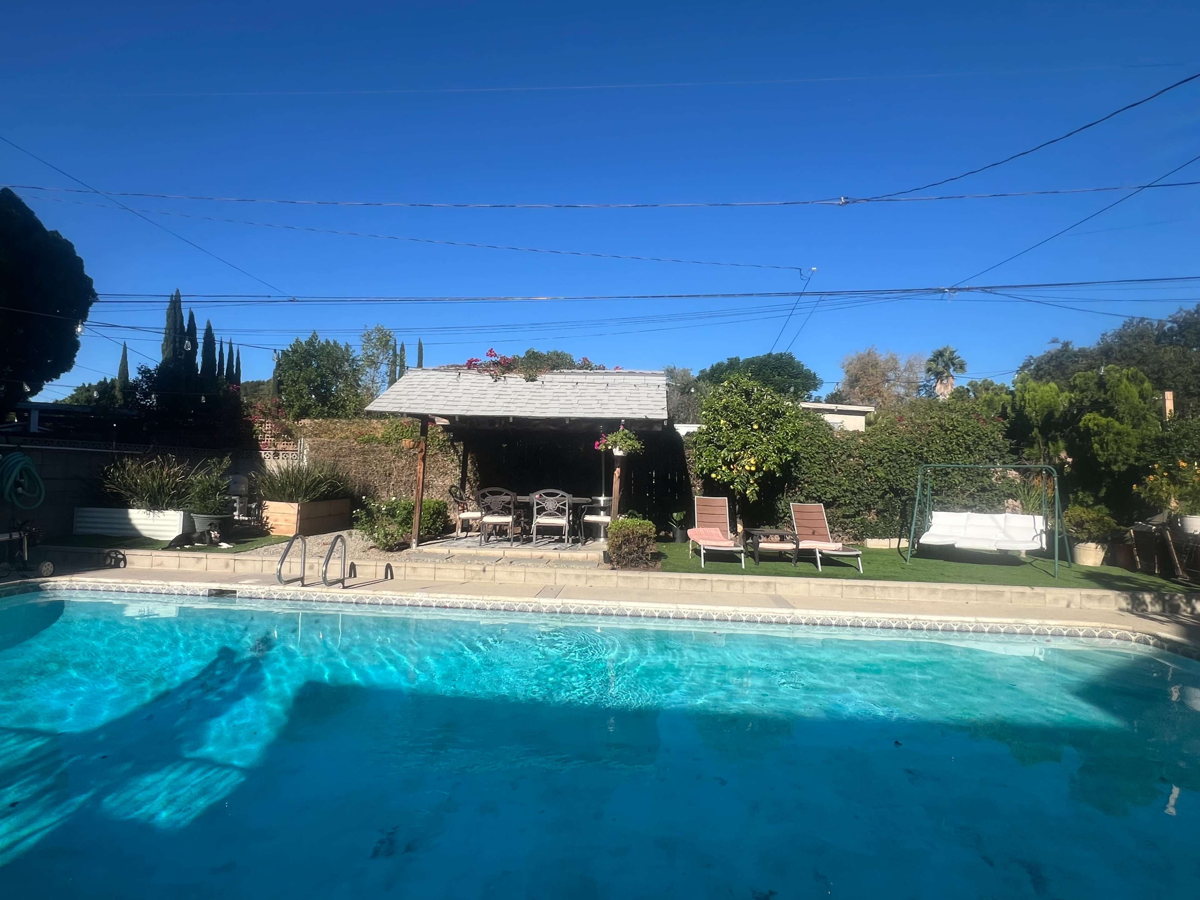 The image shows a swimming pool surrounded by lounge chairs and a shaded seating area with trees and plants in the background under a clear blue sky.