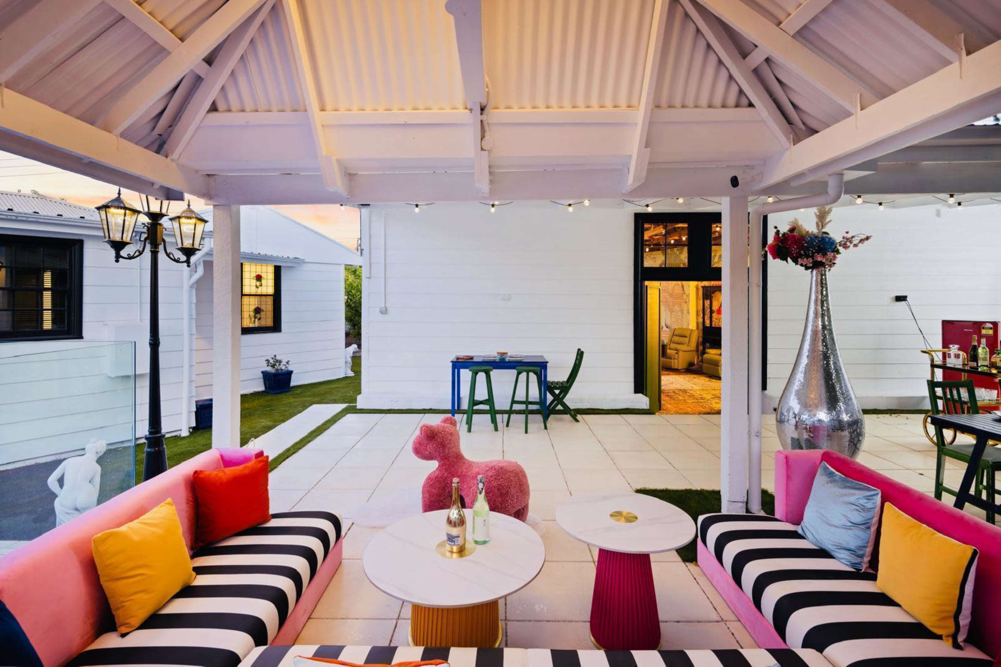 The image shows a modern outdoor lounge area with striped sofas, colorful cushions, and a pink decorative poodle, surrounded by light-colored walls and a grassy lawn.