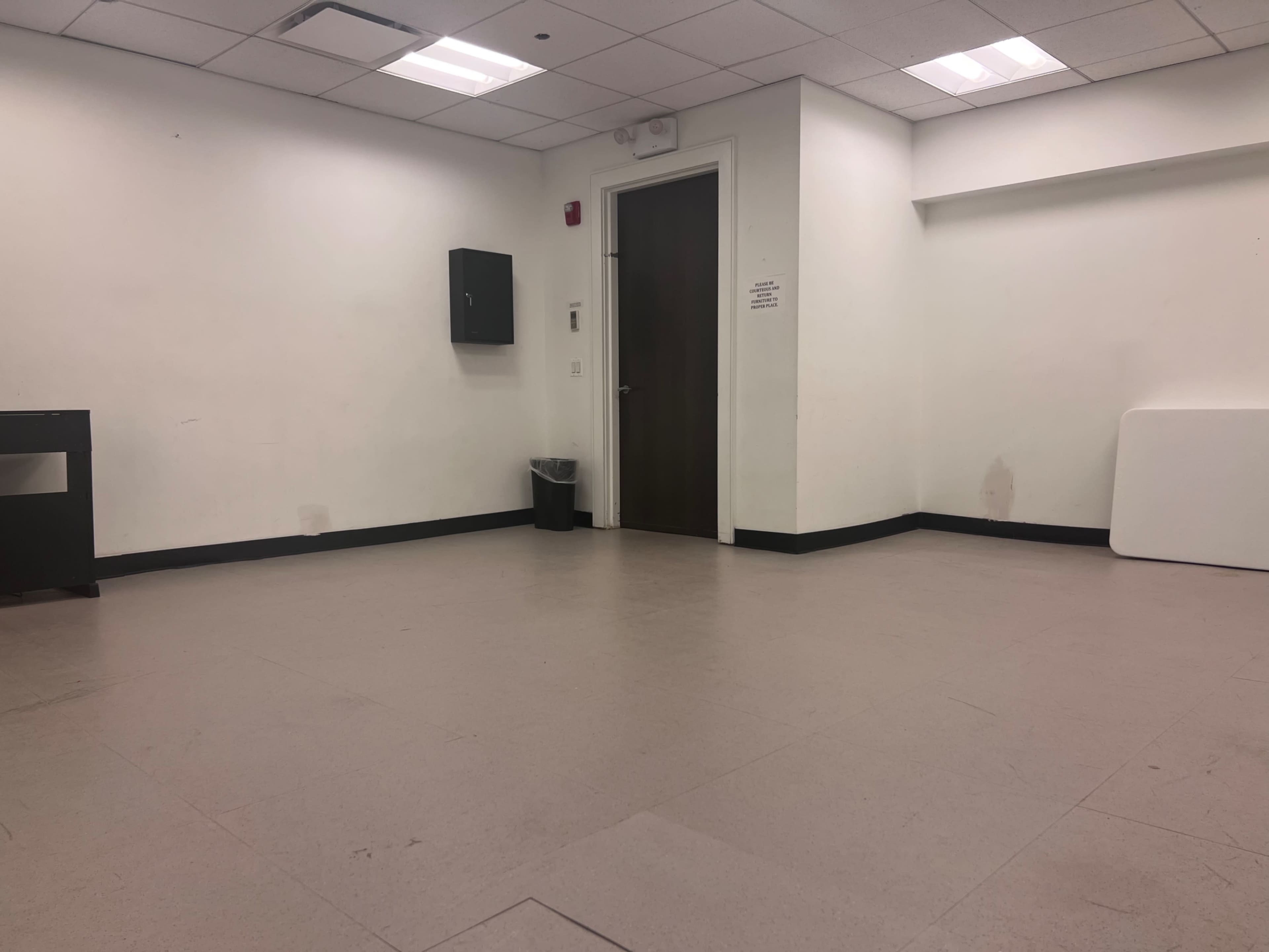 The image shows a vacant room with light-colored walls, a door at one end, a trash can, and a black cabinet, with a white object on the floor.