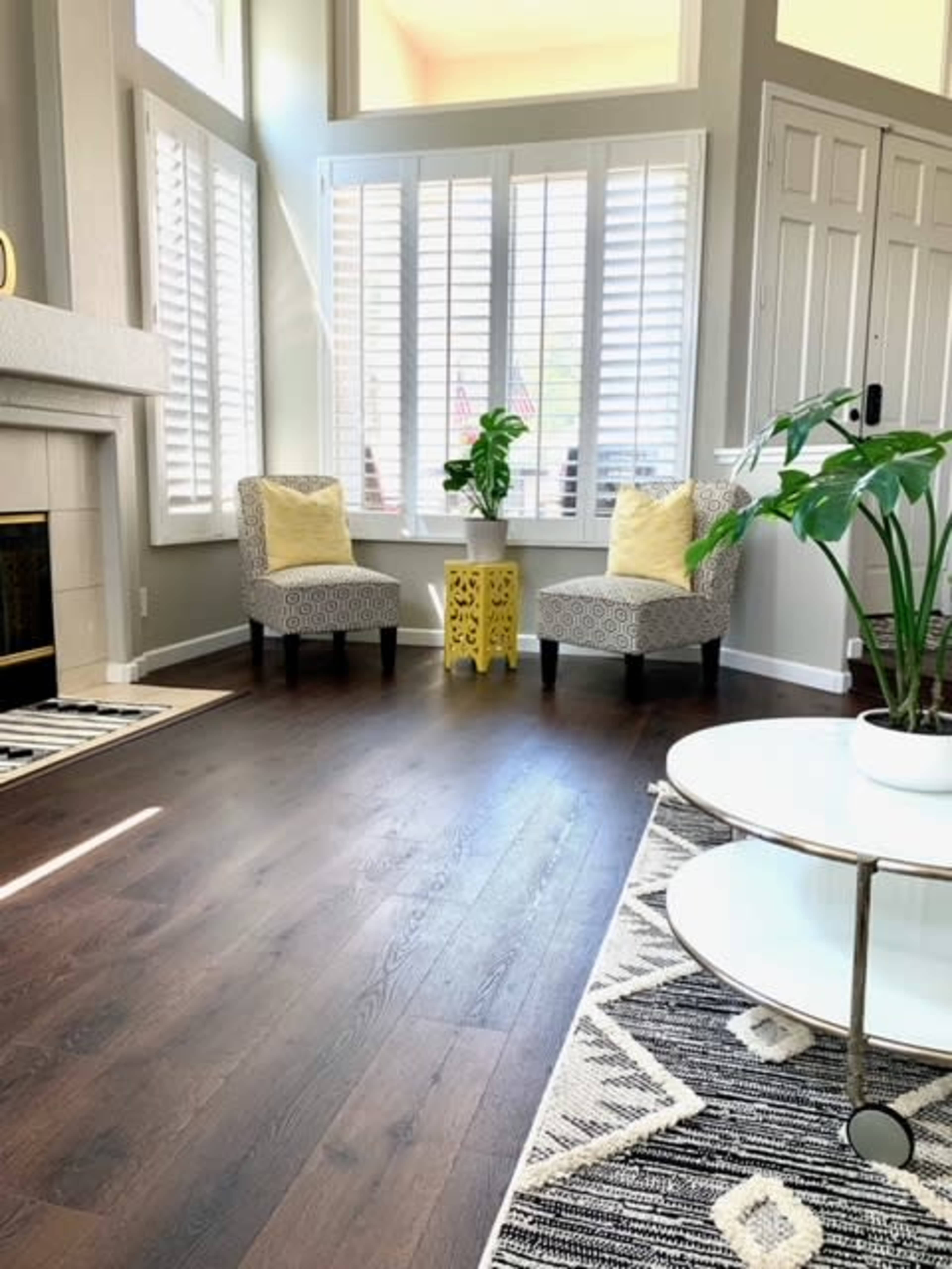 A living room with two patterned chairs, a small yellow table, a potted plant, and a fireplace, featuring large windows with white shutters.