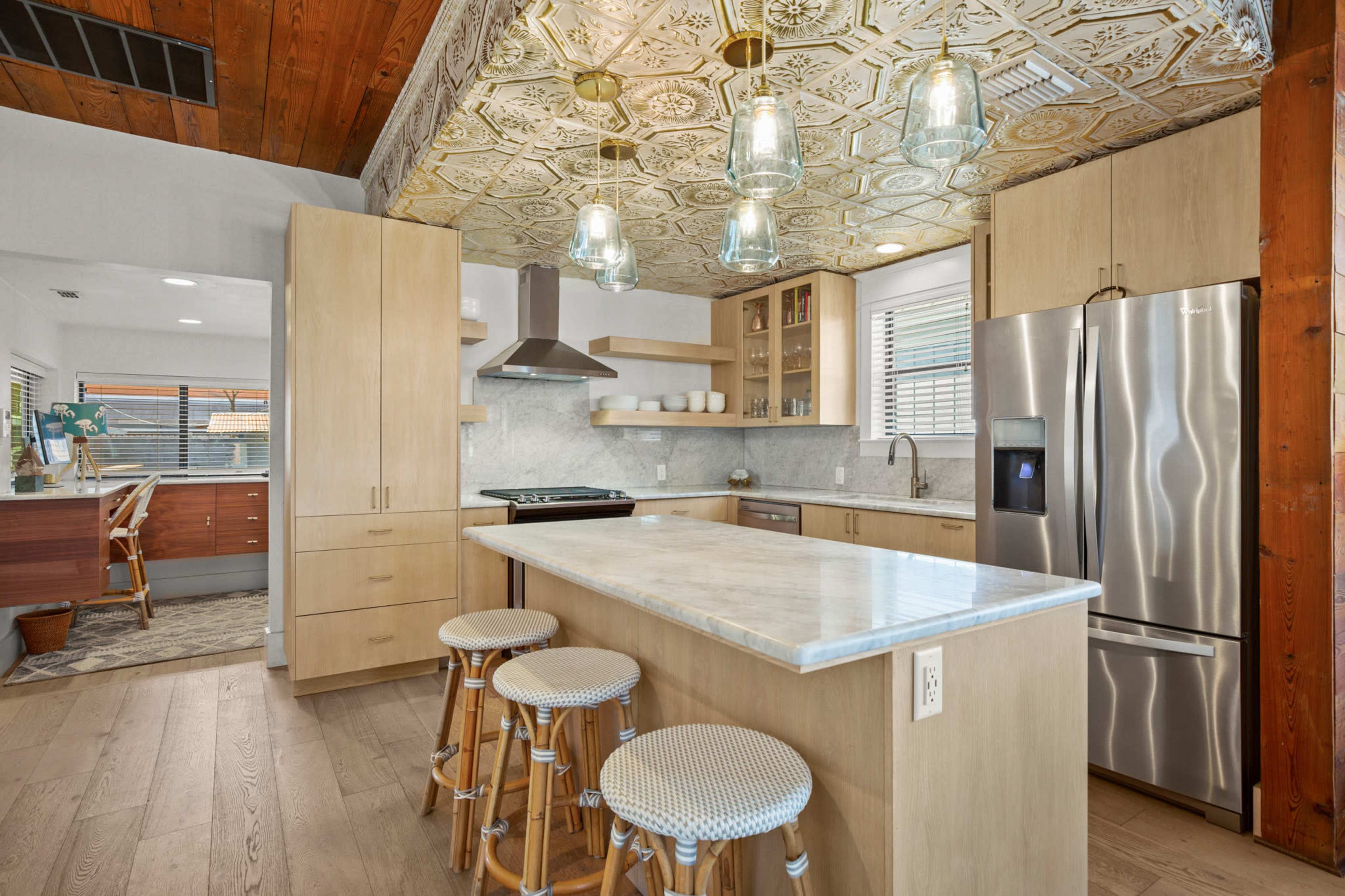 A modern kitchen features light wood cabinetry, a marble countertop island with three stools, and decorative pendant lighting above.