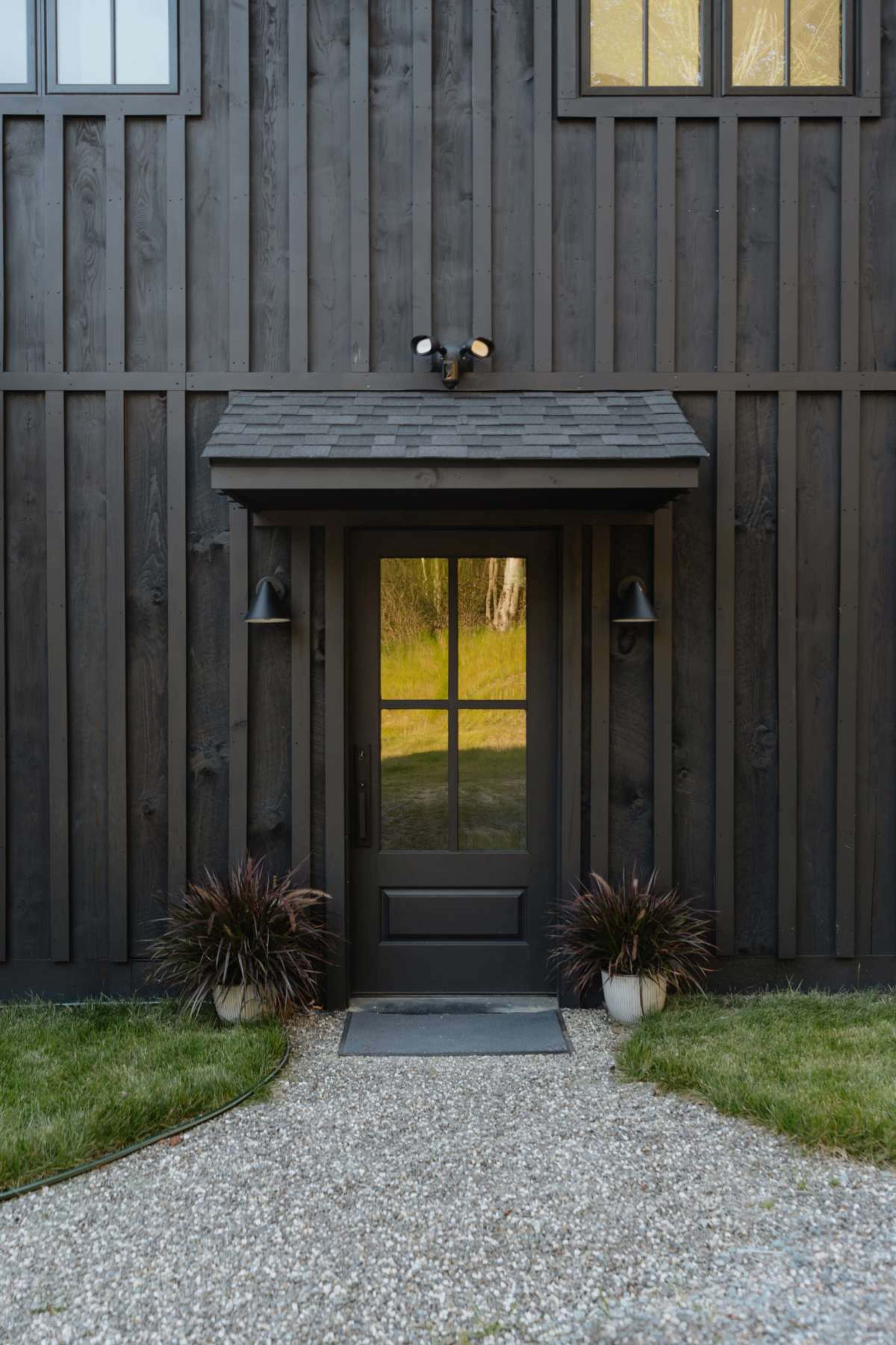 The image shows a black wood-paneled entrance with a small overhang, a glass door, and planters flanking the pathway leading to it.