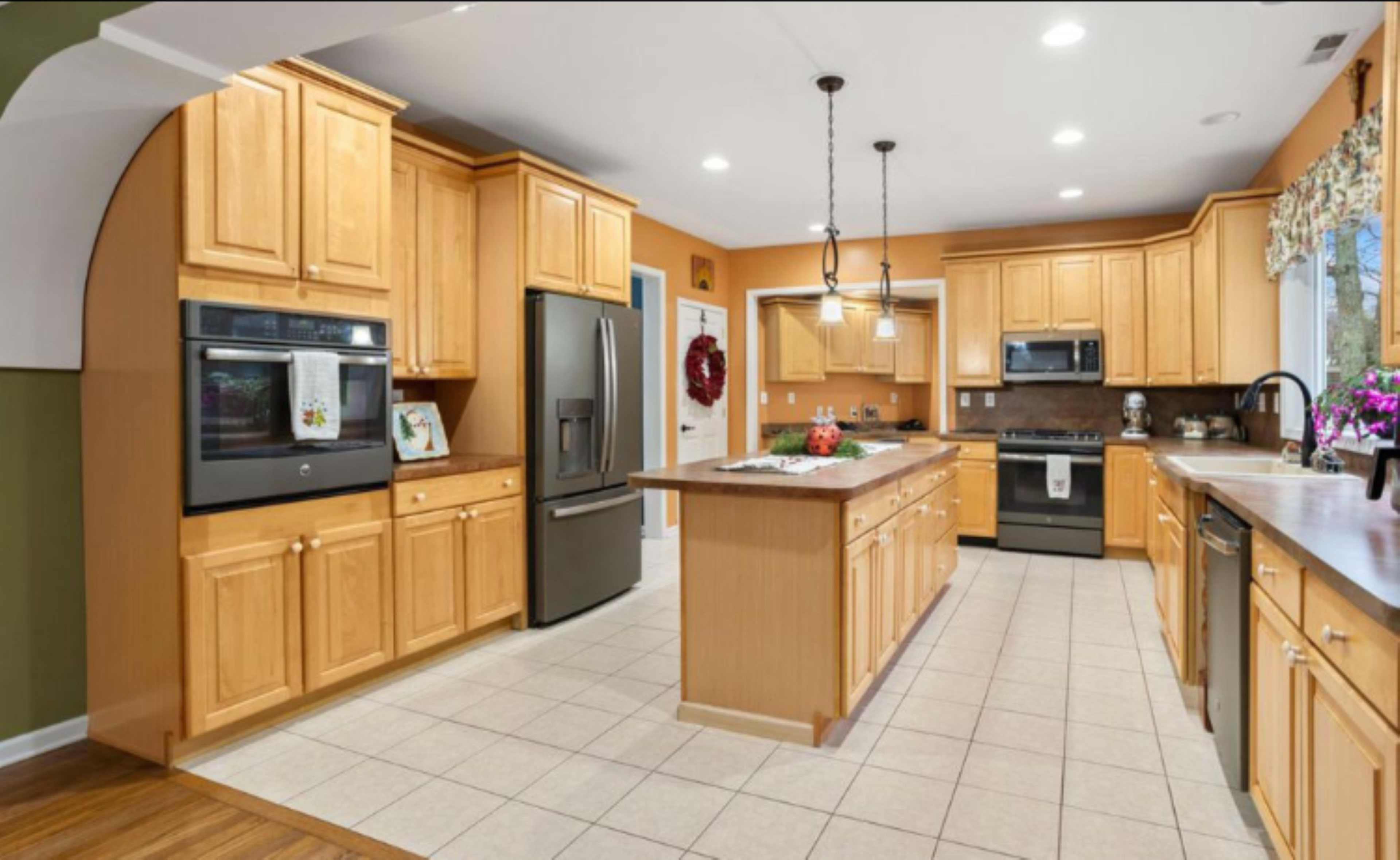 The image shows a spacious kitchen with wooden cabinets, stainless steel appliances, and a tiled floor.