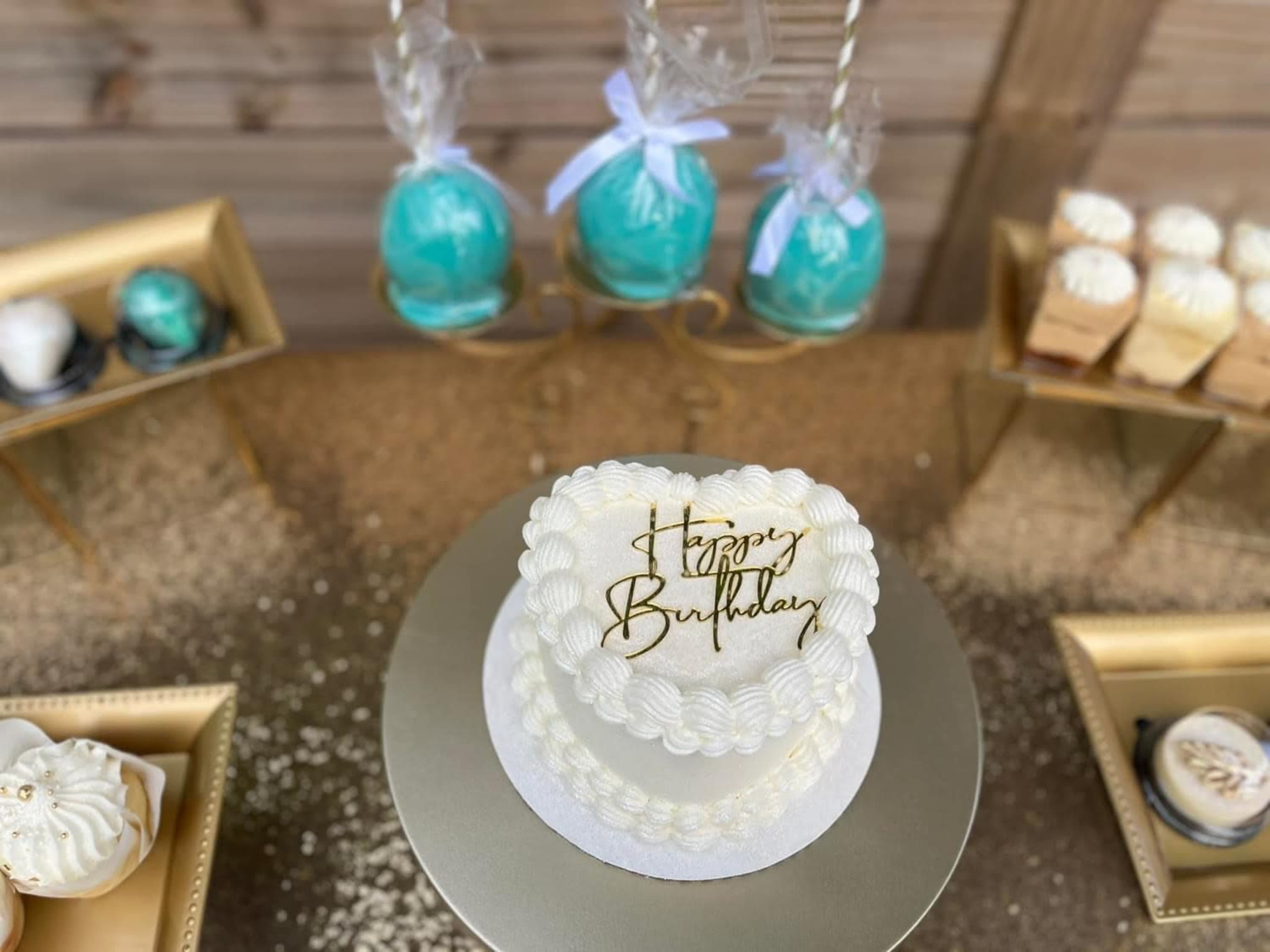 A small white birthday cake with a gold "Happy Birthday" topper is displayed on a silver pedestal, surrounded by decorative treats on golden trays.