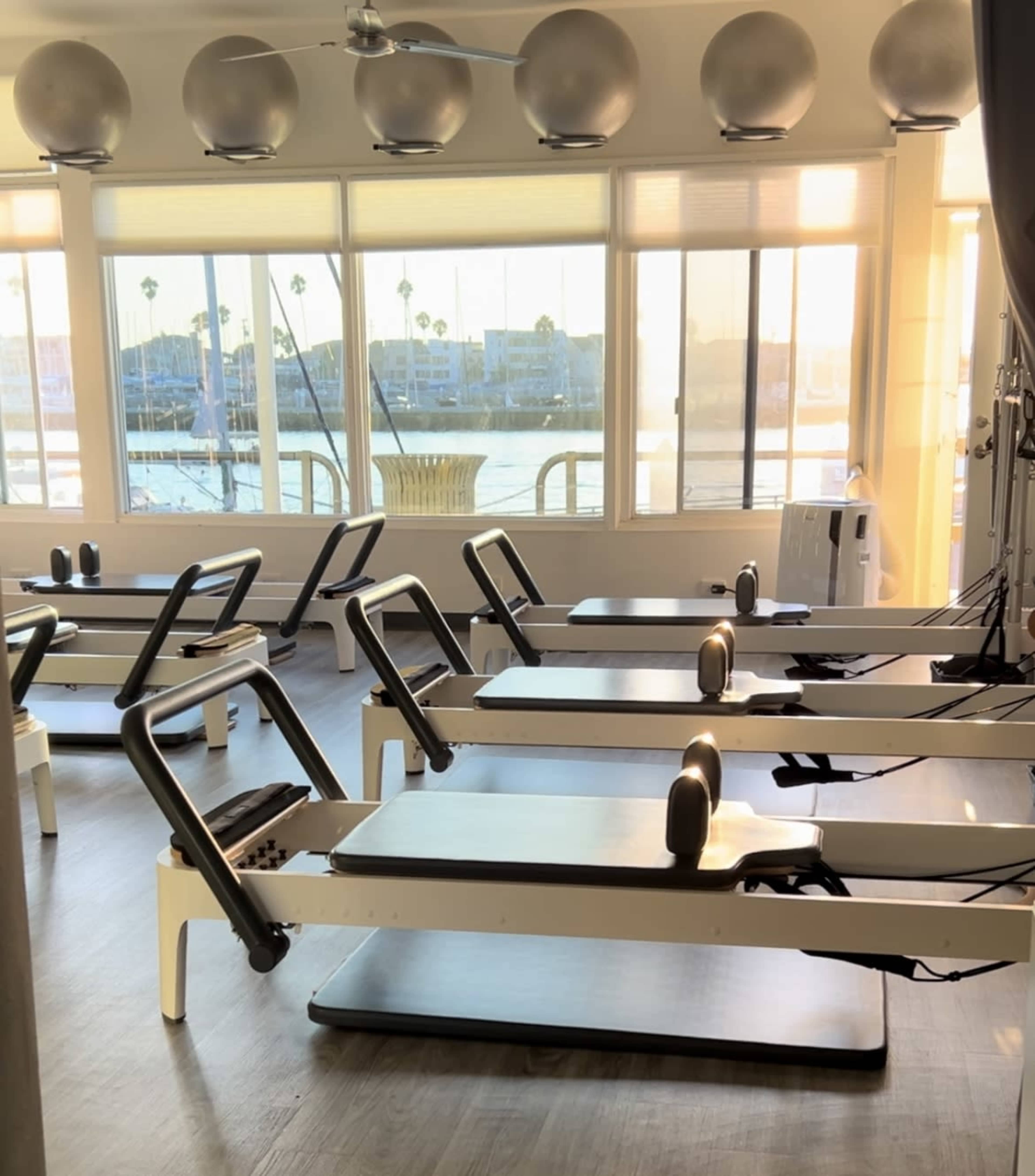 The image shows a bright fitness studio with several reformer Pilates machines arranged neatly in front of large windows overlooking a body of water.