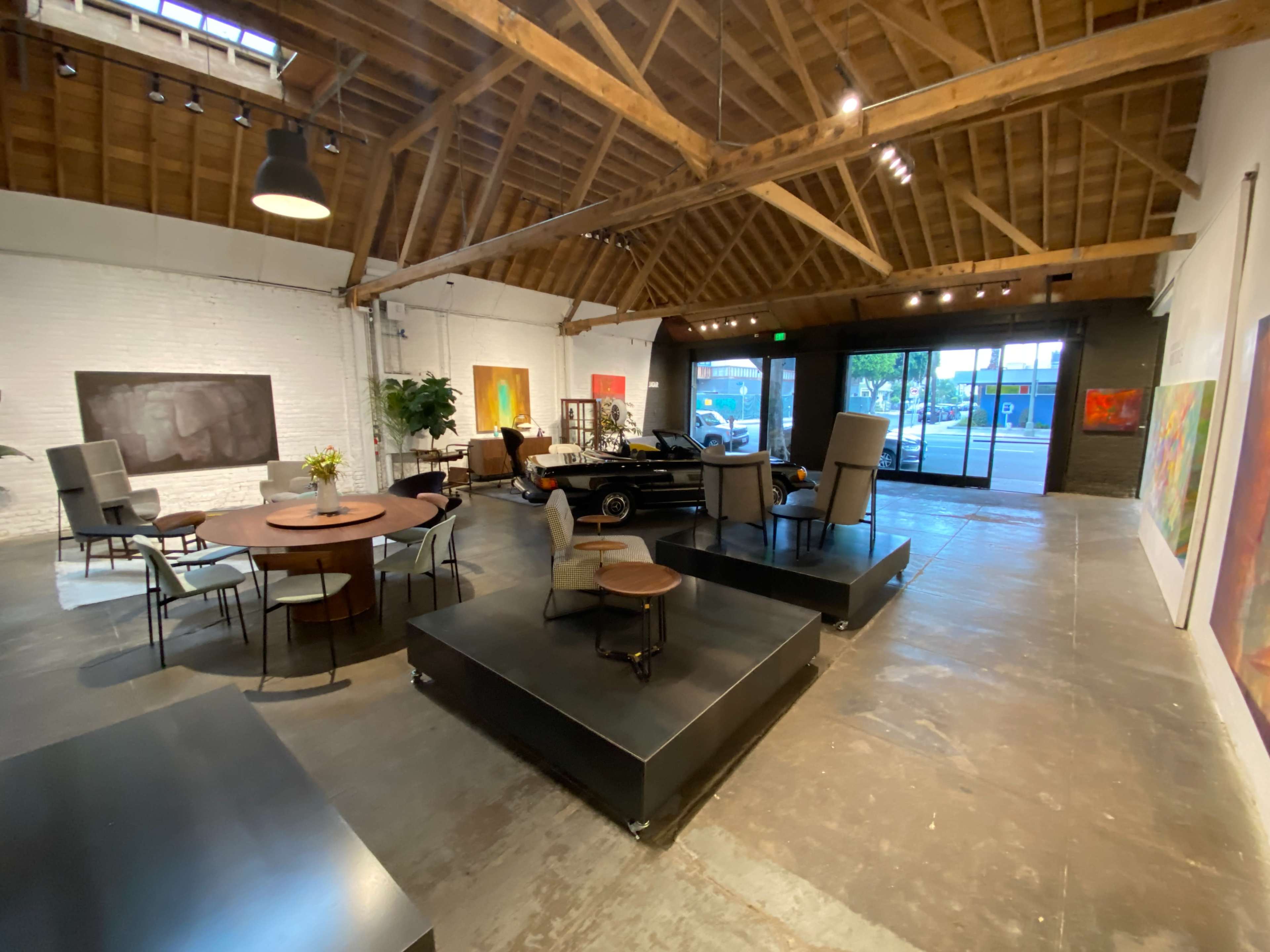 A spacious art gallery featuring various seating arrangements, a round table, and a classic car, all set under a wooden ceiling with large windows.