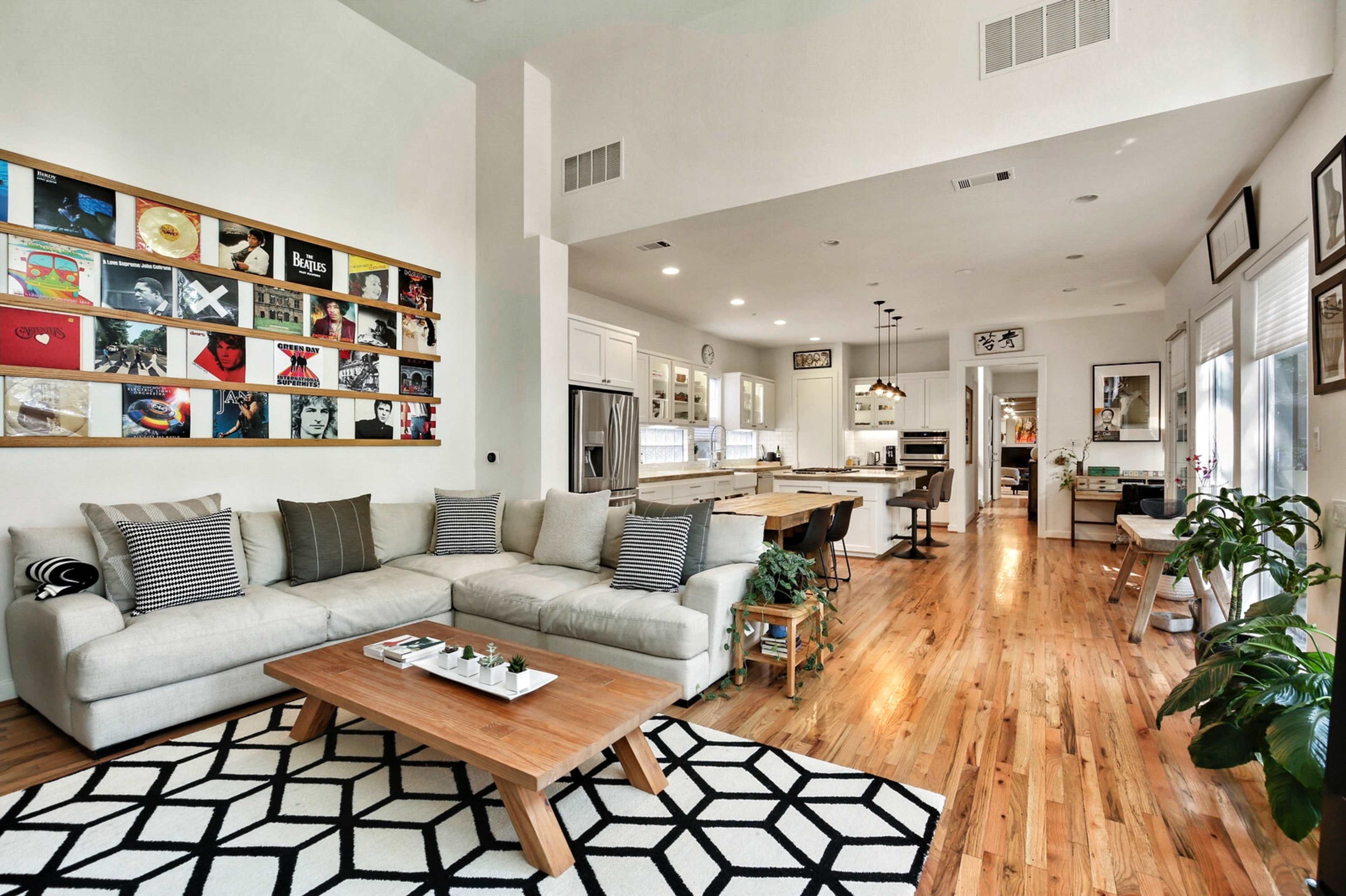 A modern living room connected to a kitchen, featuring a light-colored sectional sofa, a coffee table, and a large wall adorned with framed artwork.