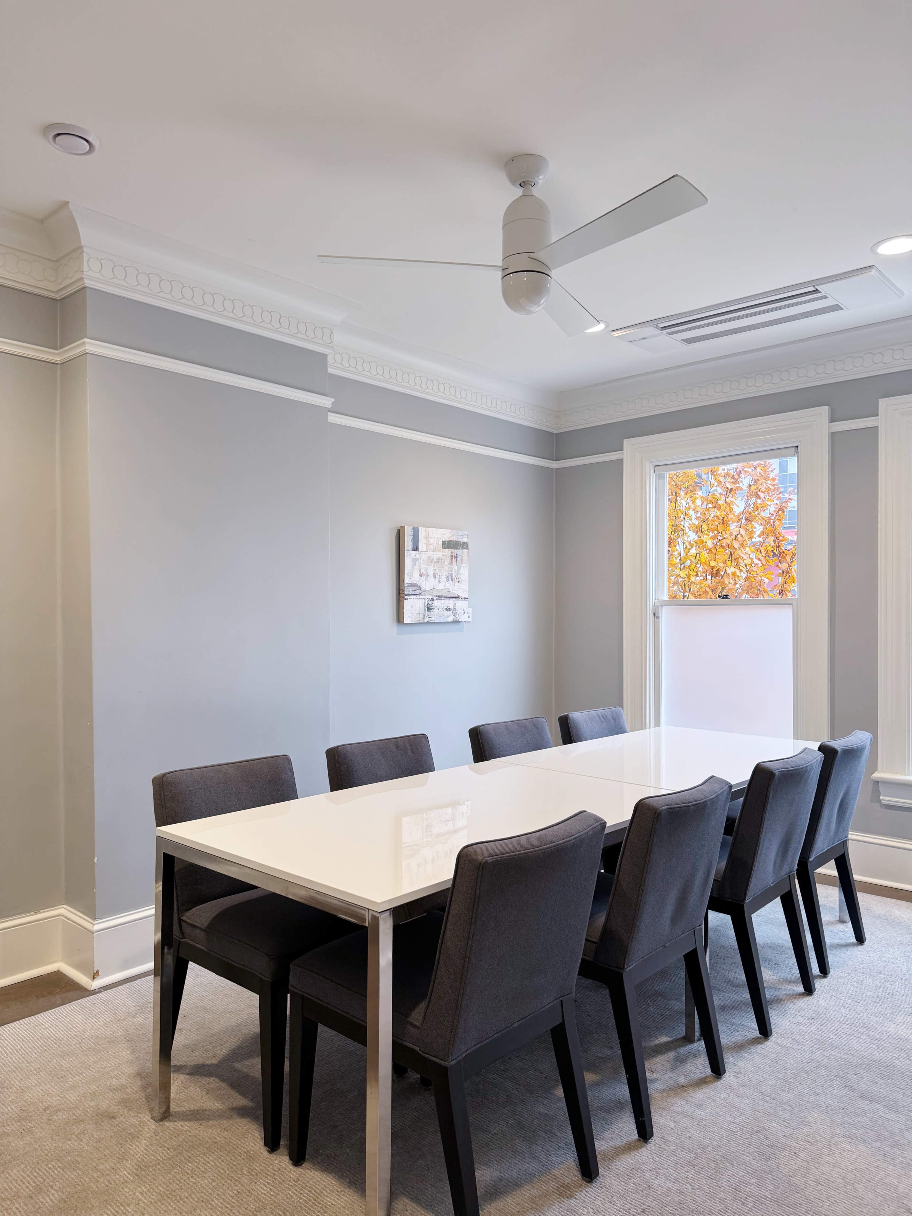 A modern conference room features a rectangular white table surrounded by eight black chairs, with a ceiling fan above and a window displaying autumn foliage outside.