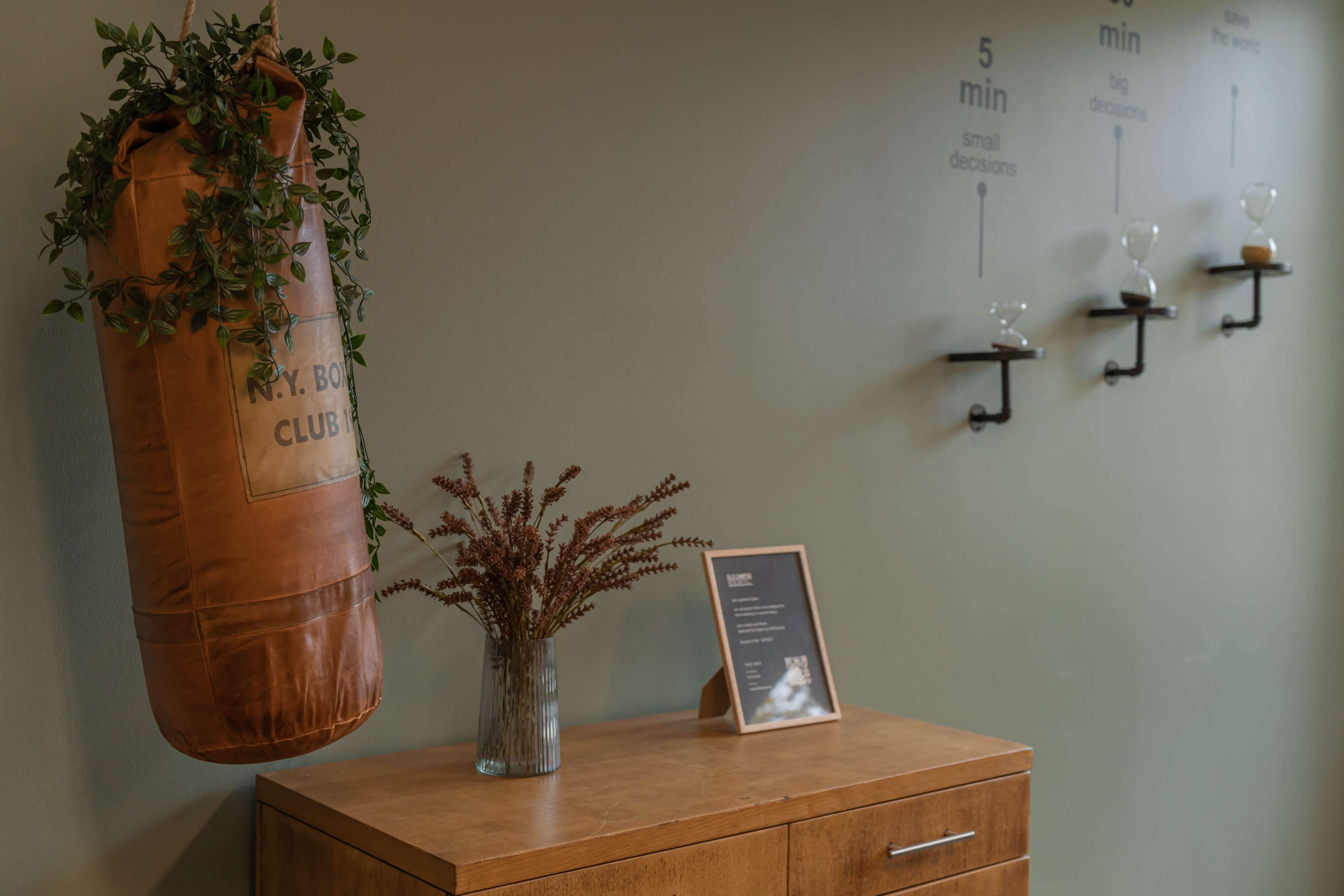 A leather punching bag hangs next to a wooden cabinet and a framed sign, with a vase of plants on top and wall-mounted shelves displaying small glass items.