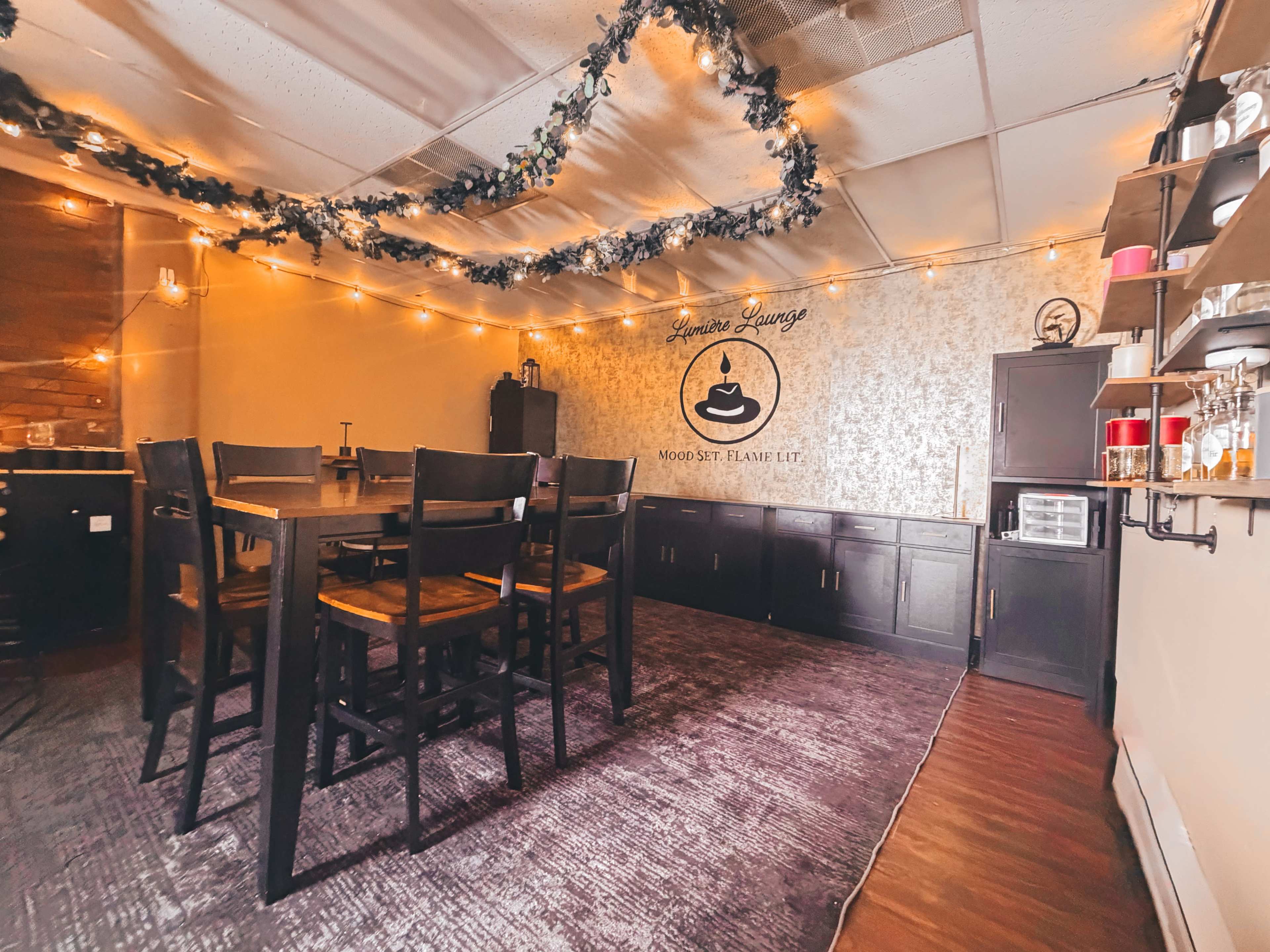 The image shows a cozy lounge area featuring a large wooden table surrounded by high chairs, with string lights and decorative garlands above.