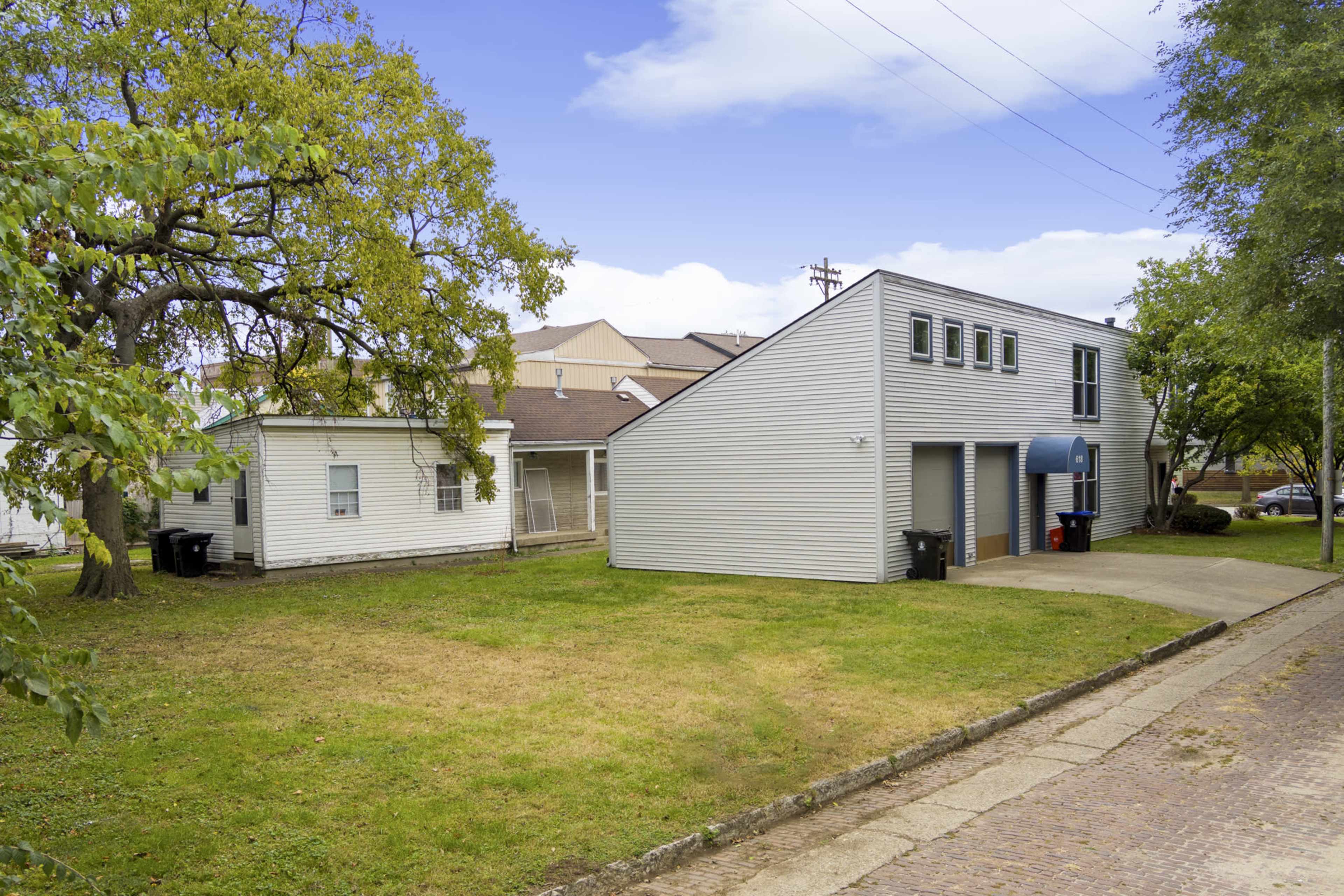 Two buildings, one with a sloped roof and blue awning, situated on a grassy area beside a paved driveway.