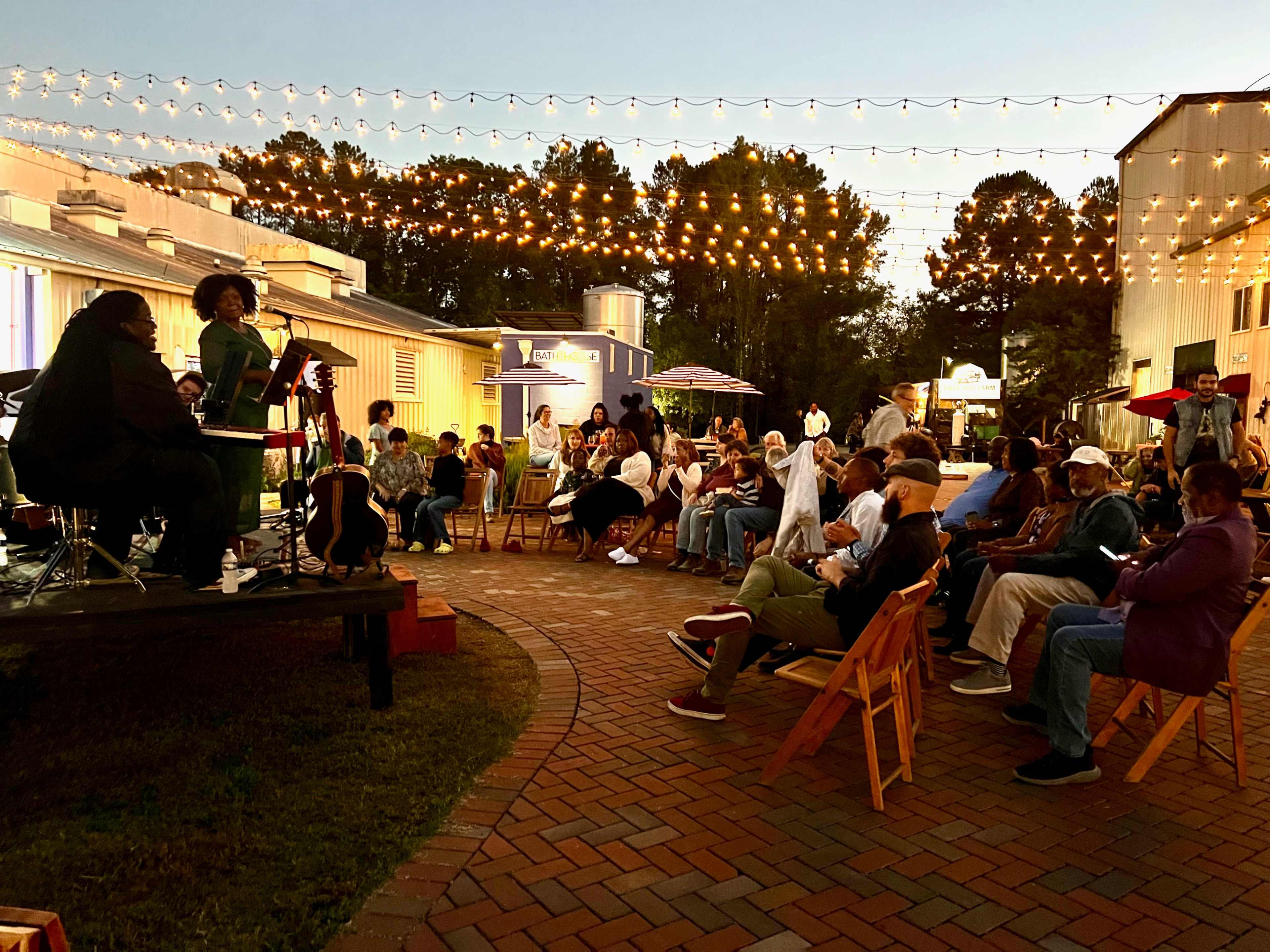 A crowd gathers in an outdoor area with string lights as a musician performs on a small stage.