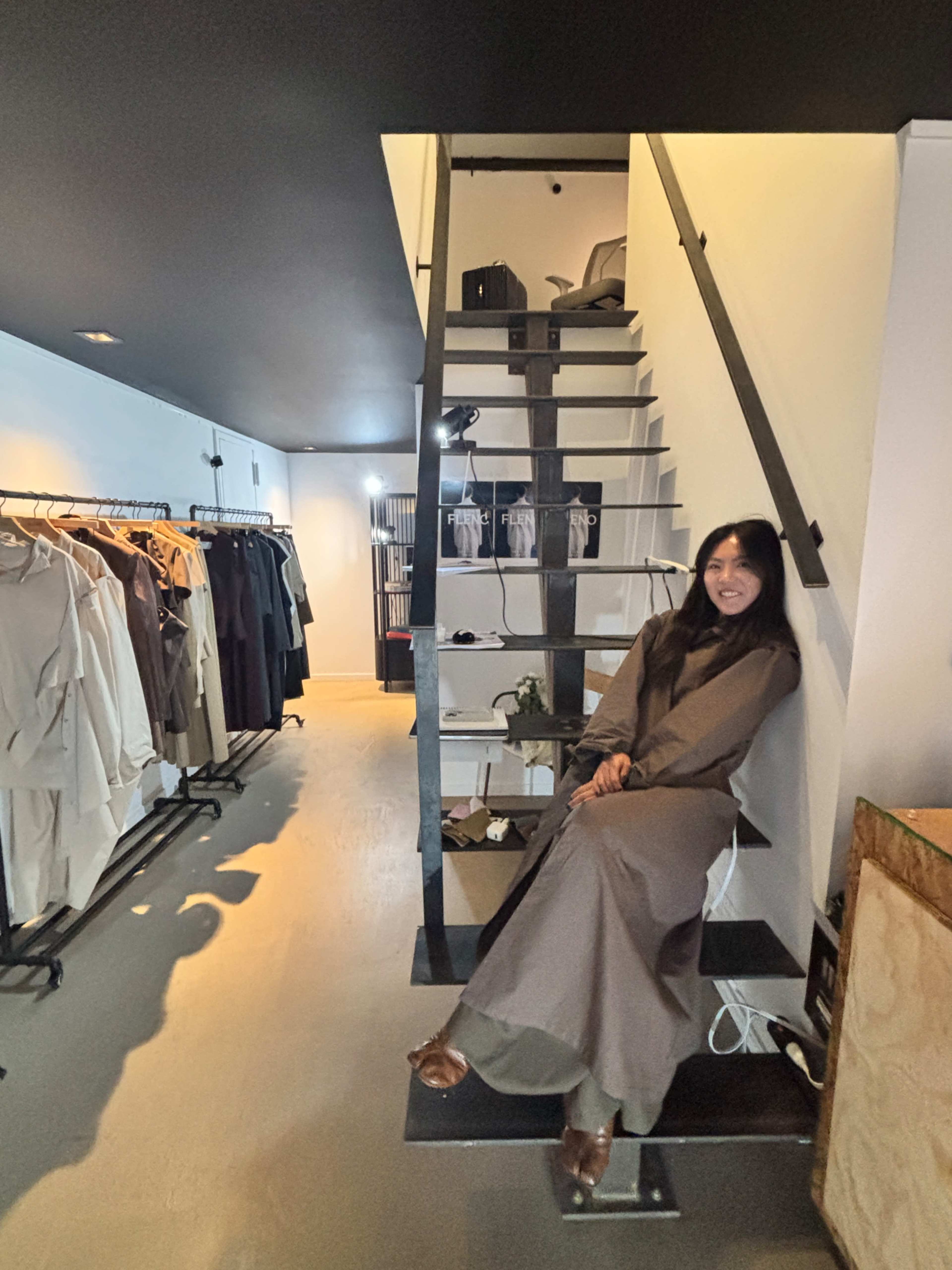 A woman sits on a metal platform beside a staircase in a clothing boutique, surrounded by racks of garments.