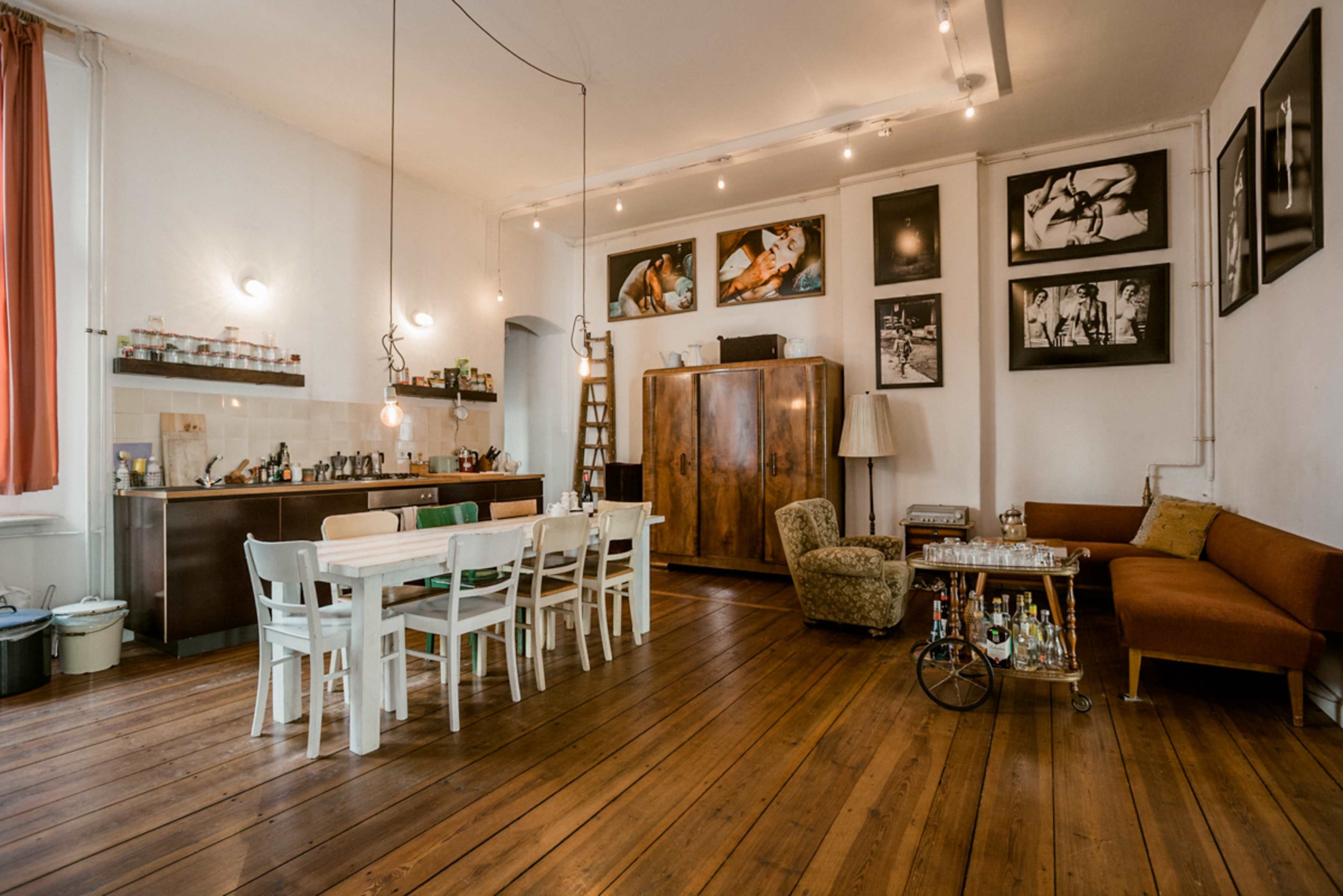 The image shows a spacious room with a kitchen area, a dining table, and a seating area, featuring wooden floors and various framed photographs on the walls.