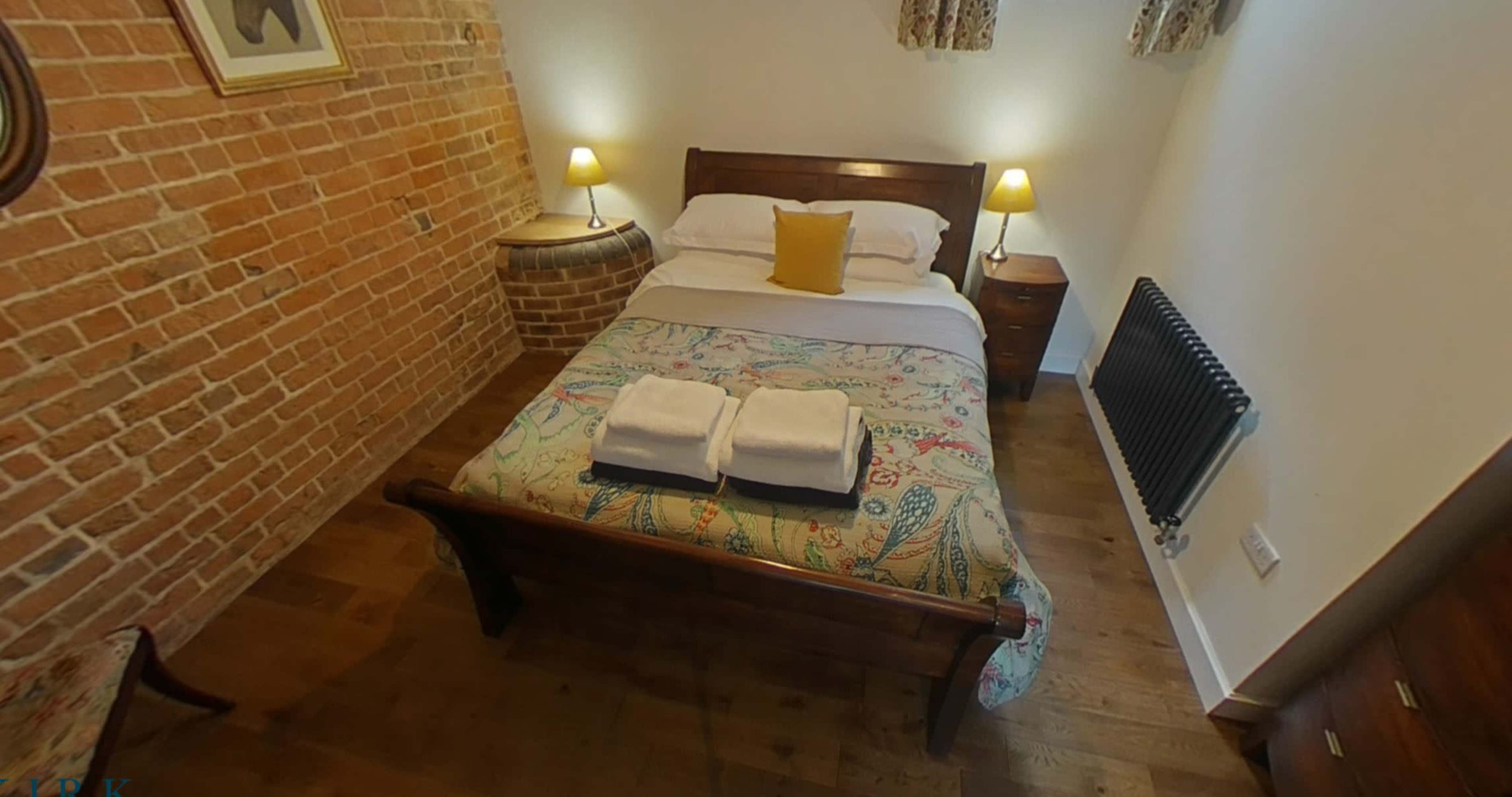 A small bedroom featuring a wooden bed with a patterned bedspread, two nightstands with lamps, and a brick wall.