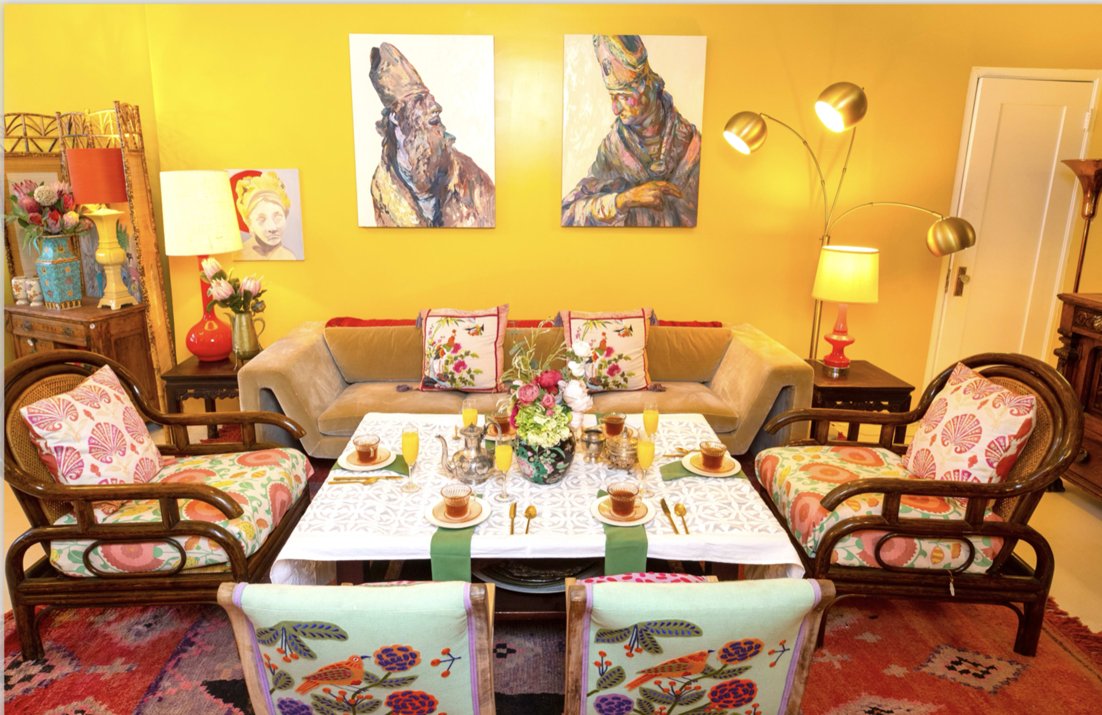 The living room features a beige sofa with patterned cushions, flanked by wooden armchairs, a table set with tea and pastries, and vibrant artwork on the yellow walls.