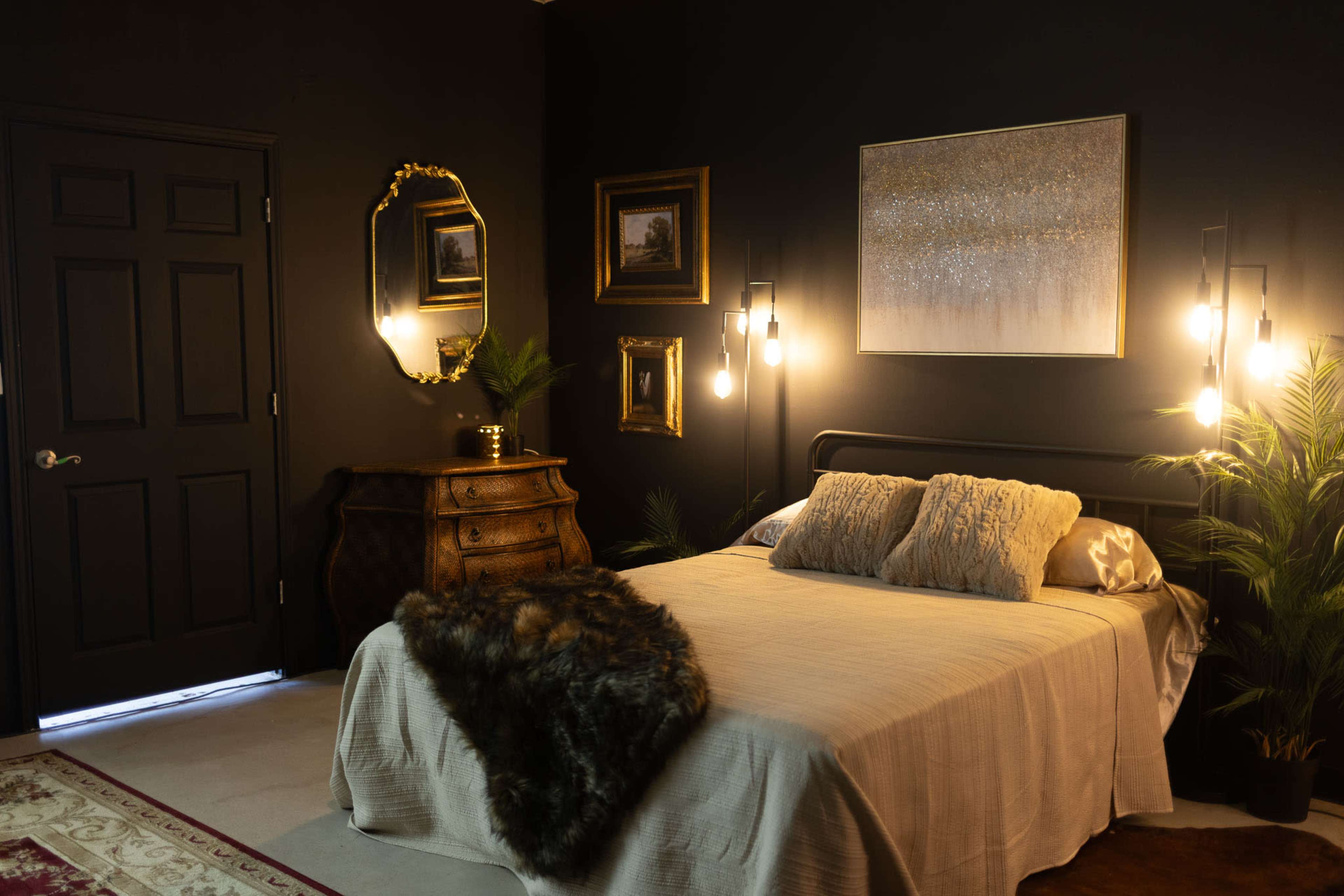 The image shows a dimly lit bedroom with a bed adorned in neutral linens, a wooden dresser, plants, and framed artworks on dark walls.