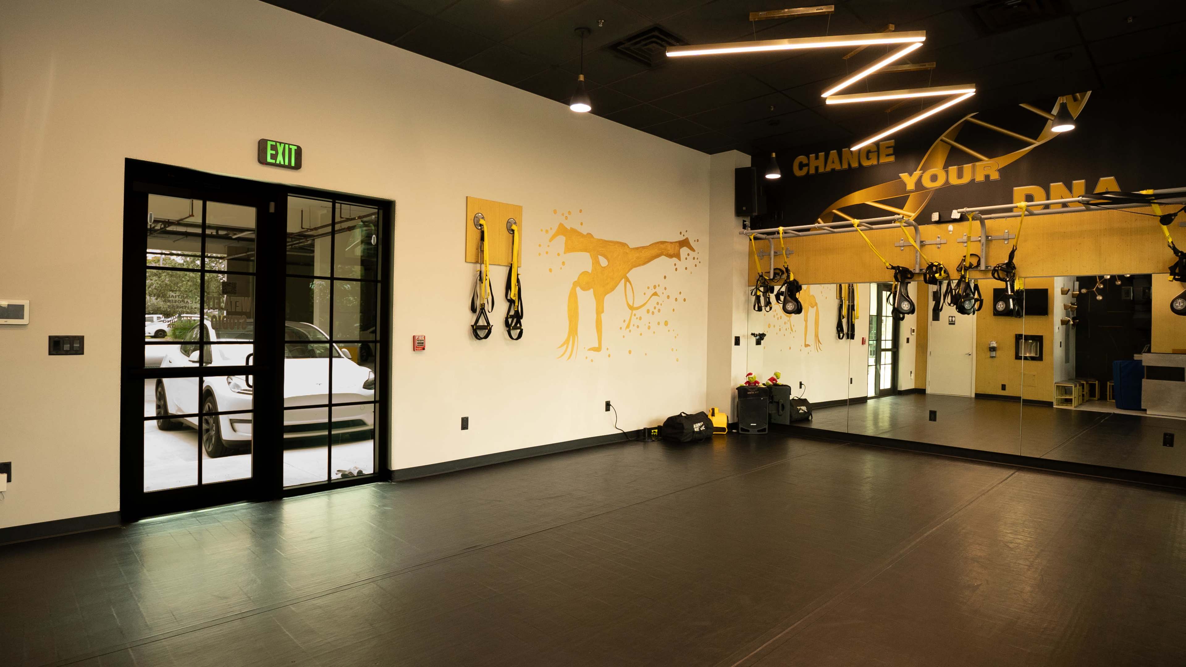 The image shows a fitness studio featuring a mirrored wall with exercise equipment mounted on the ceiling, along with a glass door leading outside.