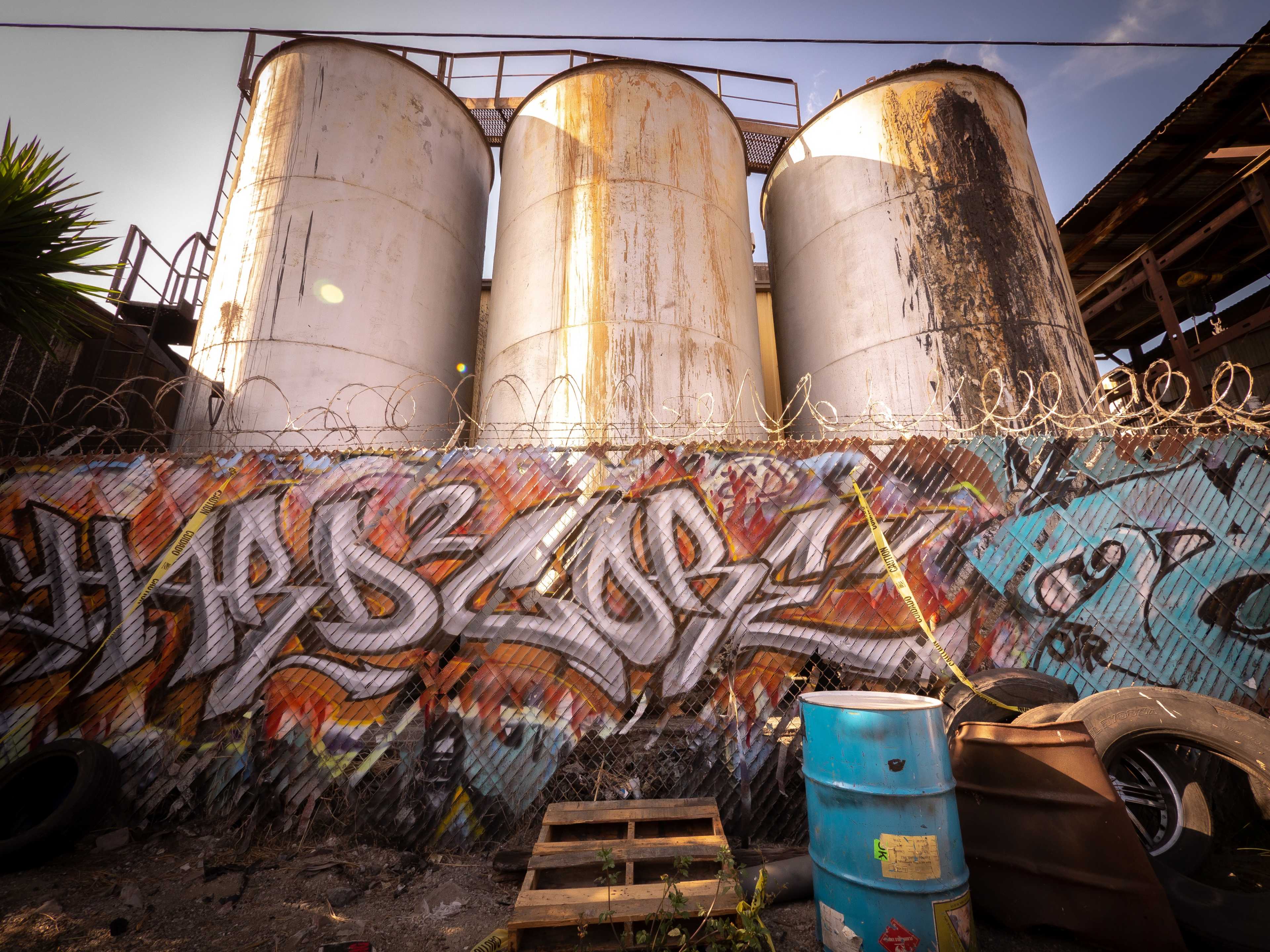 Three large metal storage tanks stand alongside a wall covered in colorful graffiti, with a barbed-wire fence in the foreground.