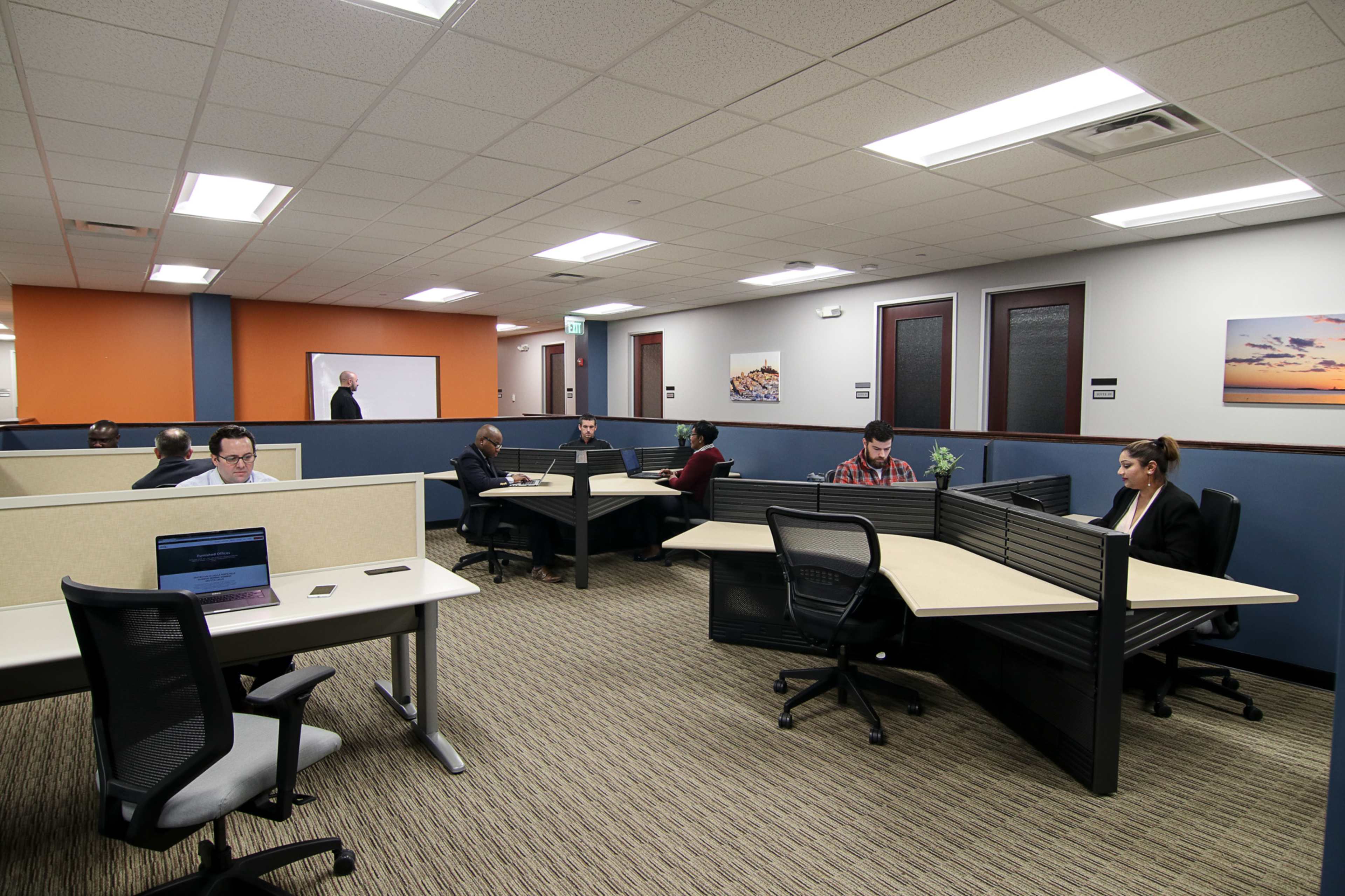 A modern office space with several workstations occupied by individuals using laptops in a well-lit environment.