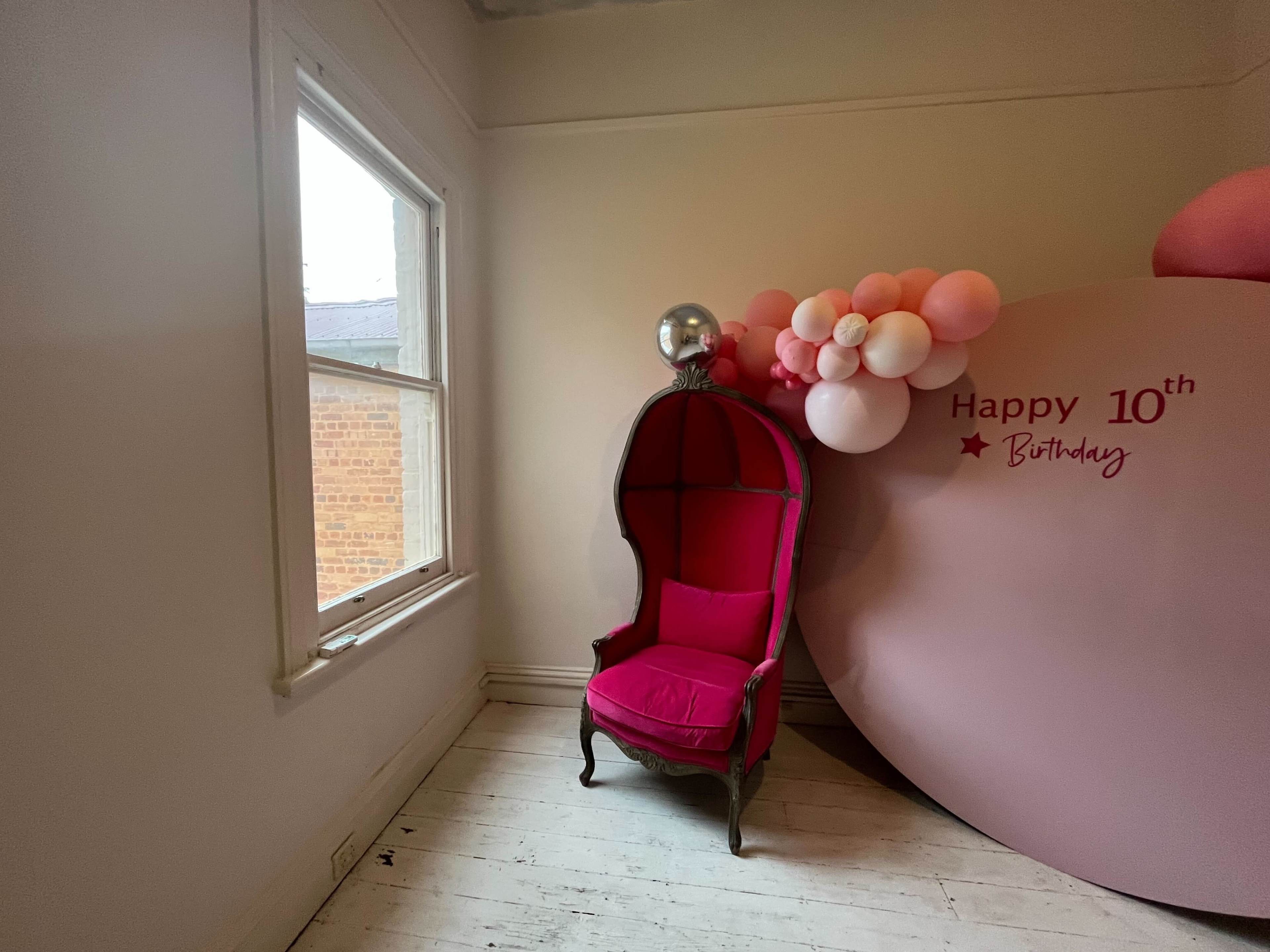 A pink chair in a room with a large round decoration that reads "Happy 10th Birthday" and a cluster of balloons nearby.