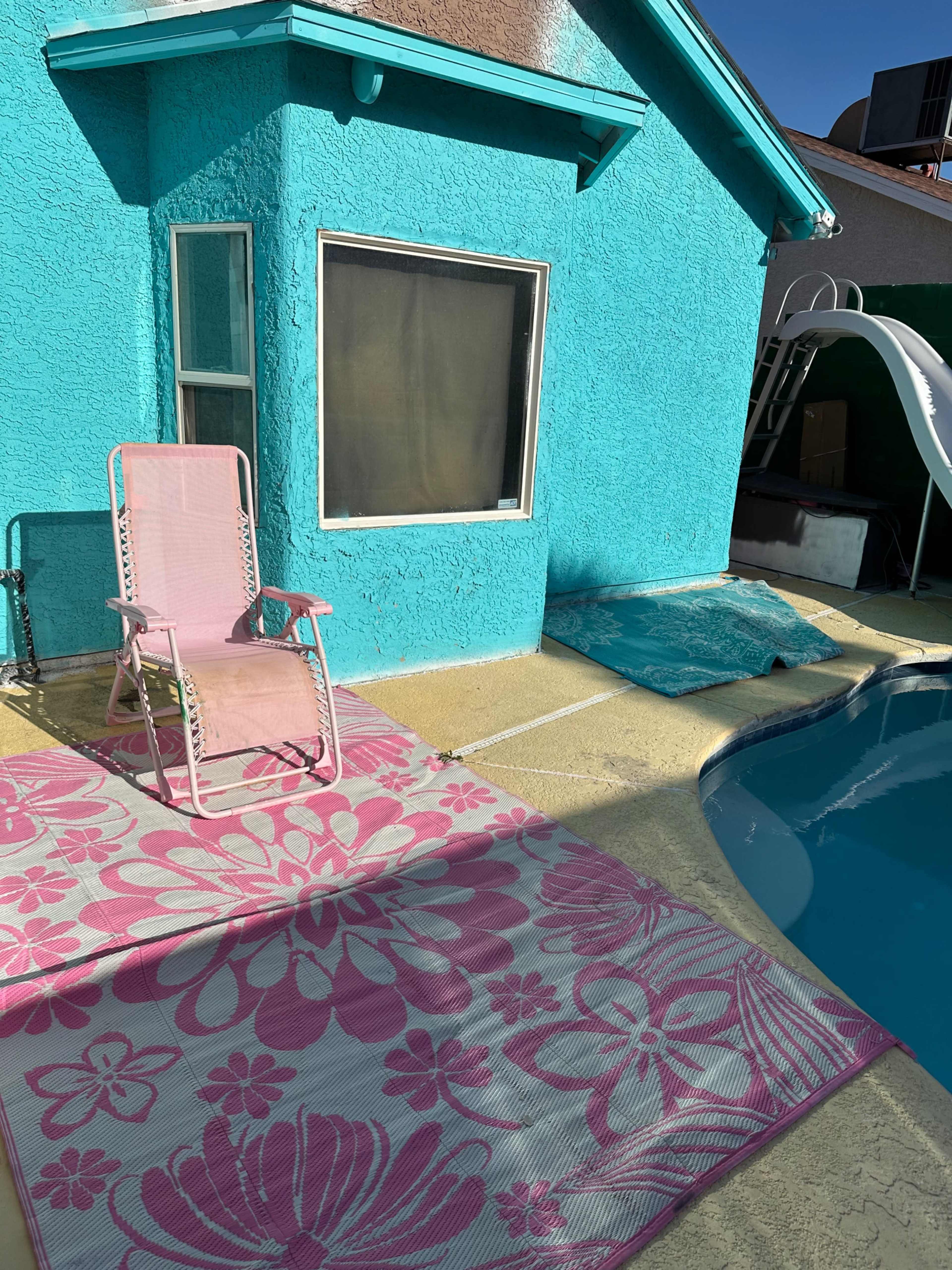 A pink chair sits on a patterned rug next to a pool beside a bright blue house.