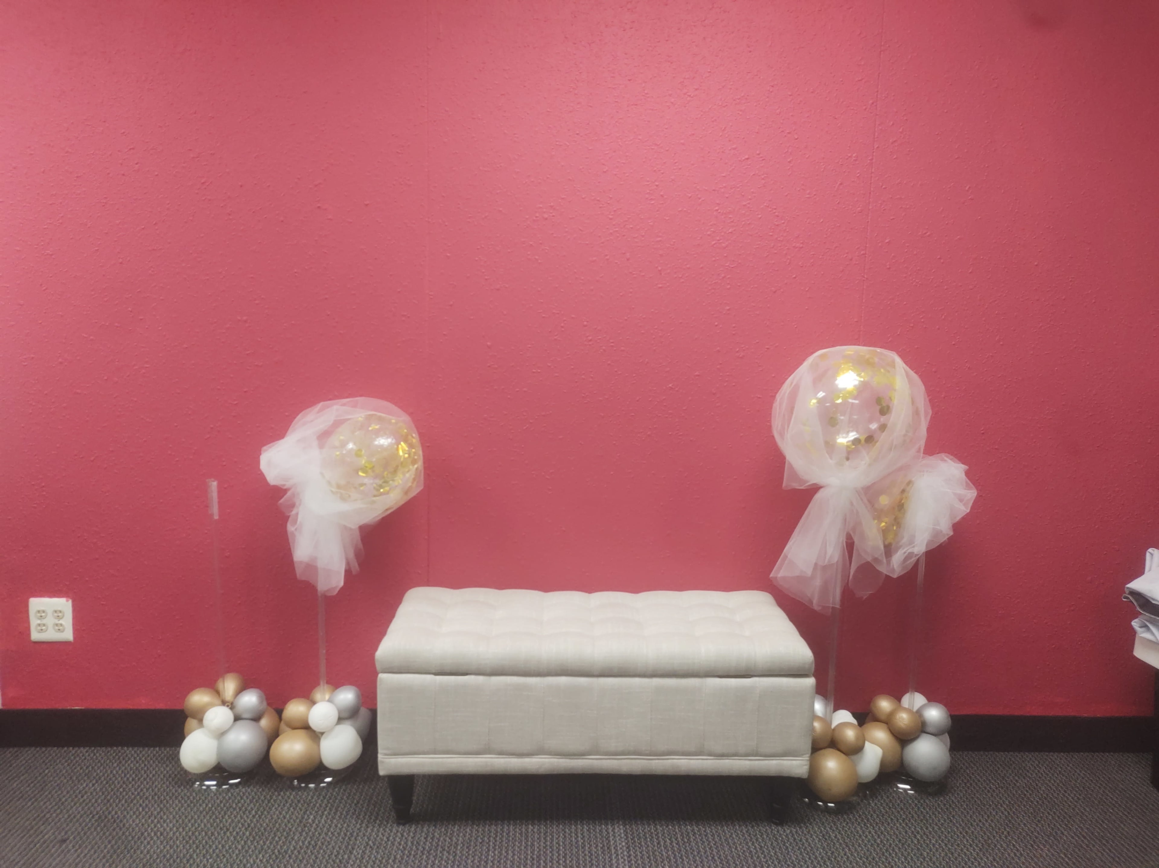 A light-colored bench in front of a pink wall, flanked by two decorative arrangements of balloons on clear stands.