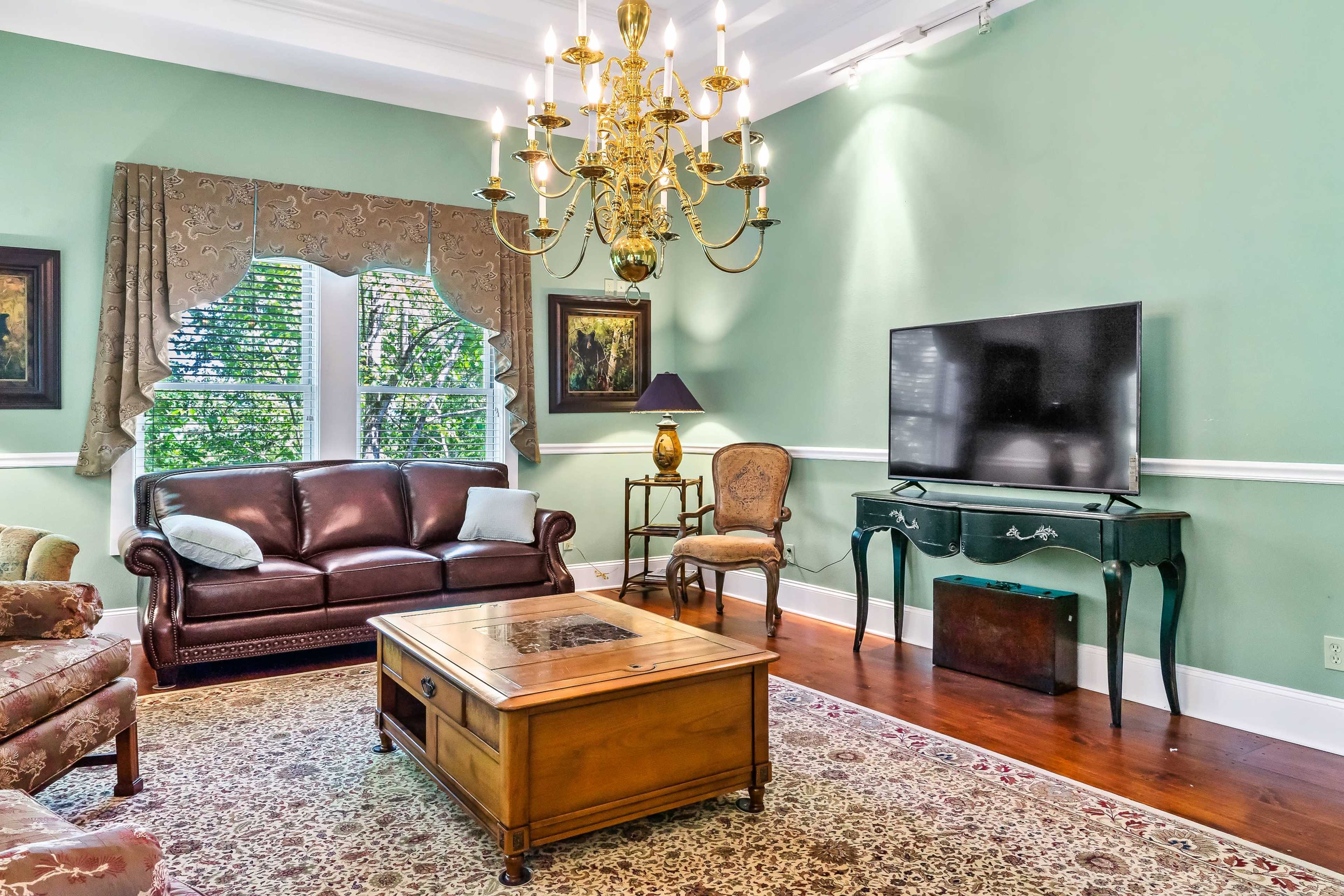 A living room with a brown leather sofa, an ornate wooden coffee table, a traditional chandelier, and a flat-screen TV on a decorative console.