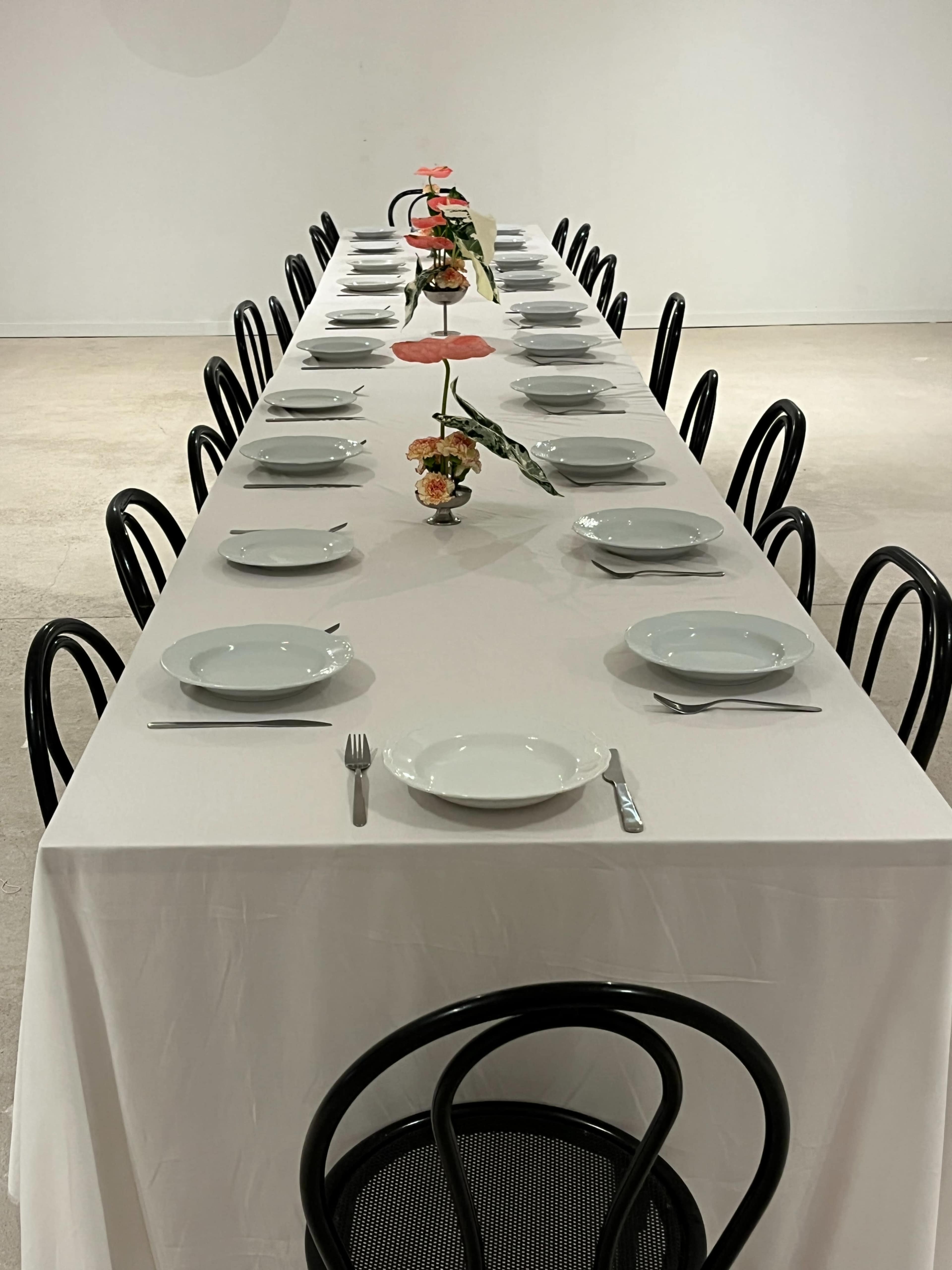 A long, rectangular dining table is set with white plates and utensils, surrounded by black chairs, with a floral centerpiece in the middle.