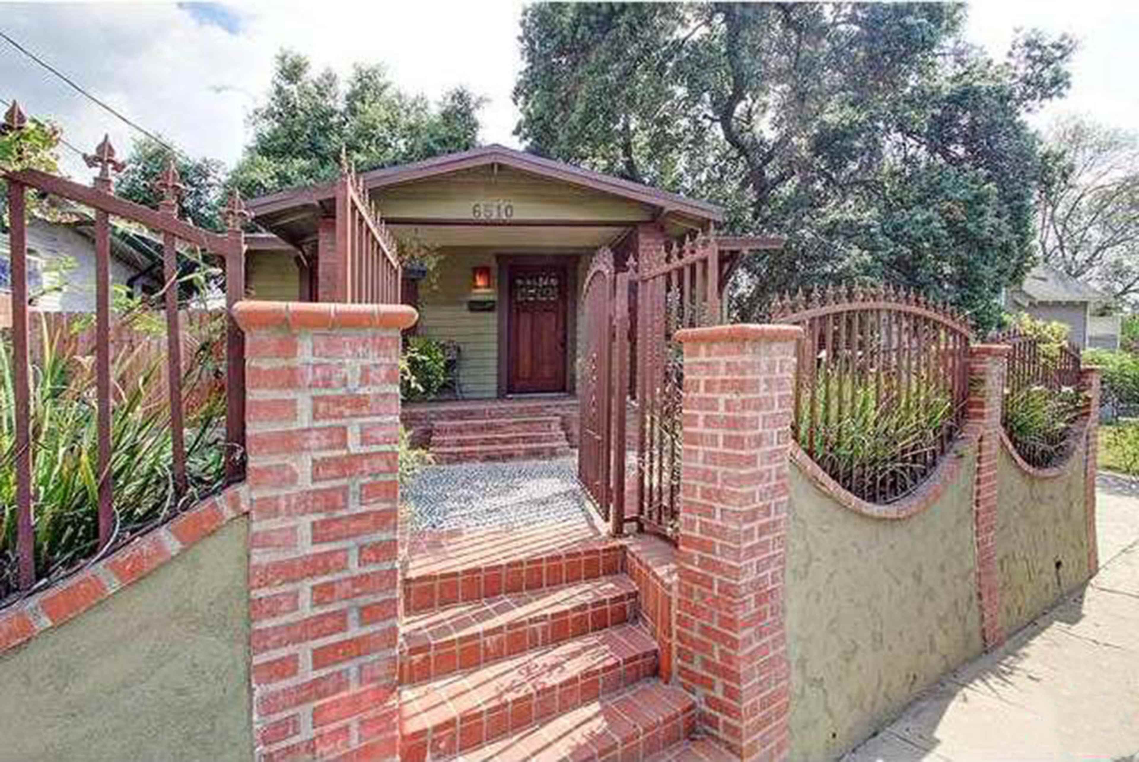 A small house with a brick walkway and decorative fencing is set behind a low wall and surrounded by greenery.