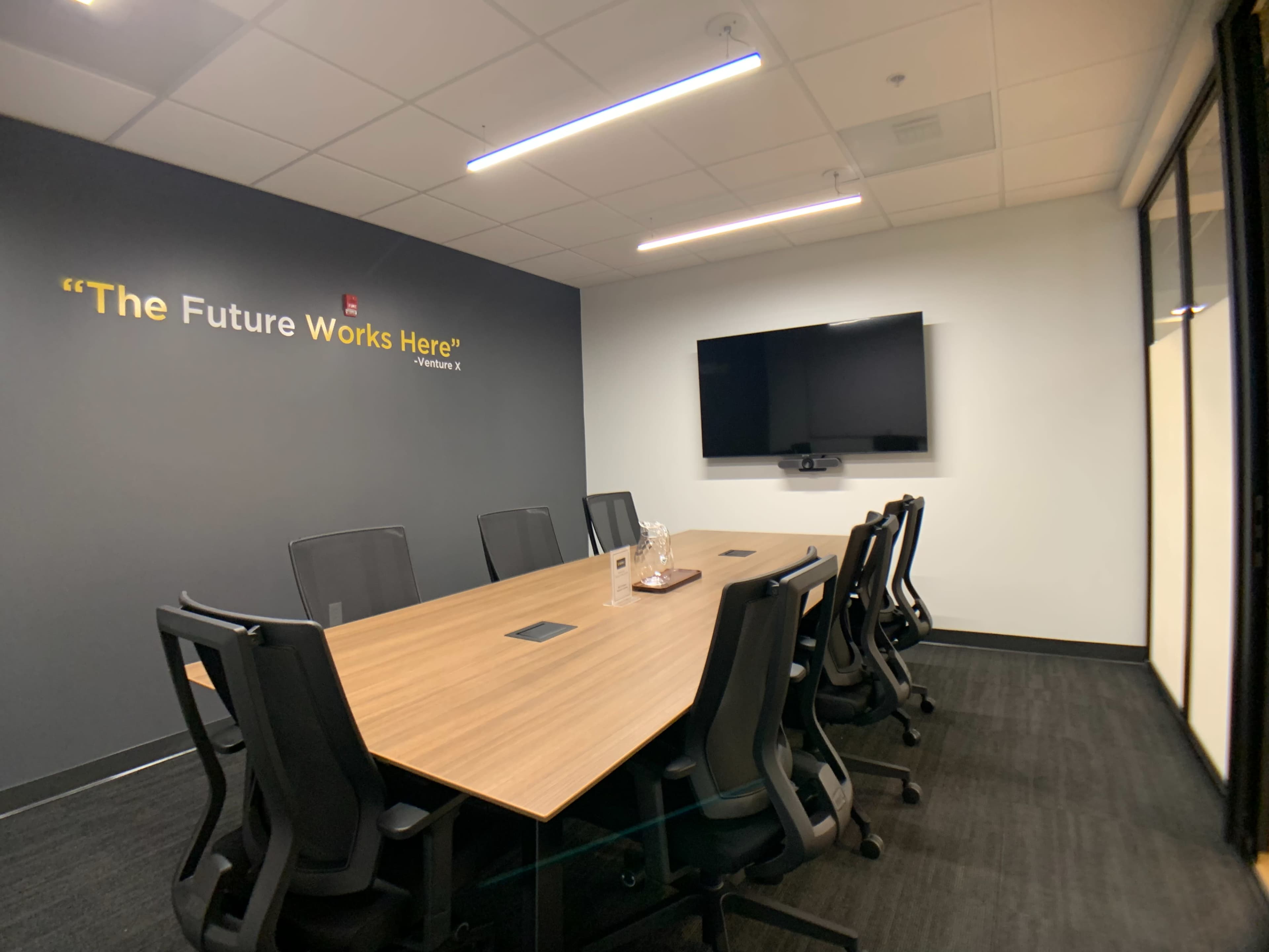 A modern conference room features a large wooden table surrounded by black chairs, a mounted television, and a wall with the phrase "The Future Works Here" above it.