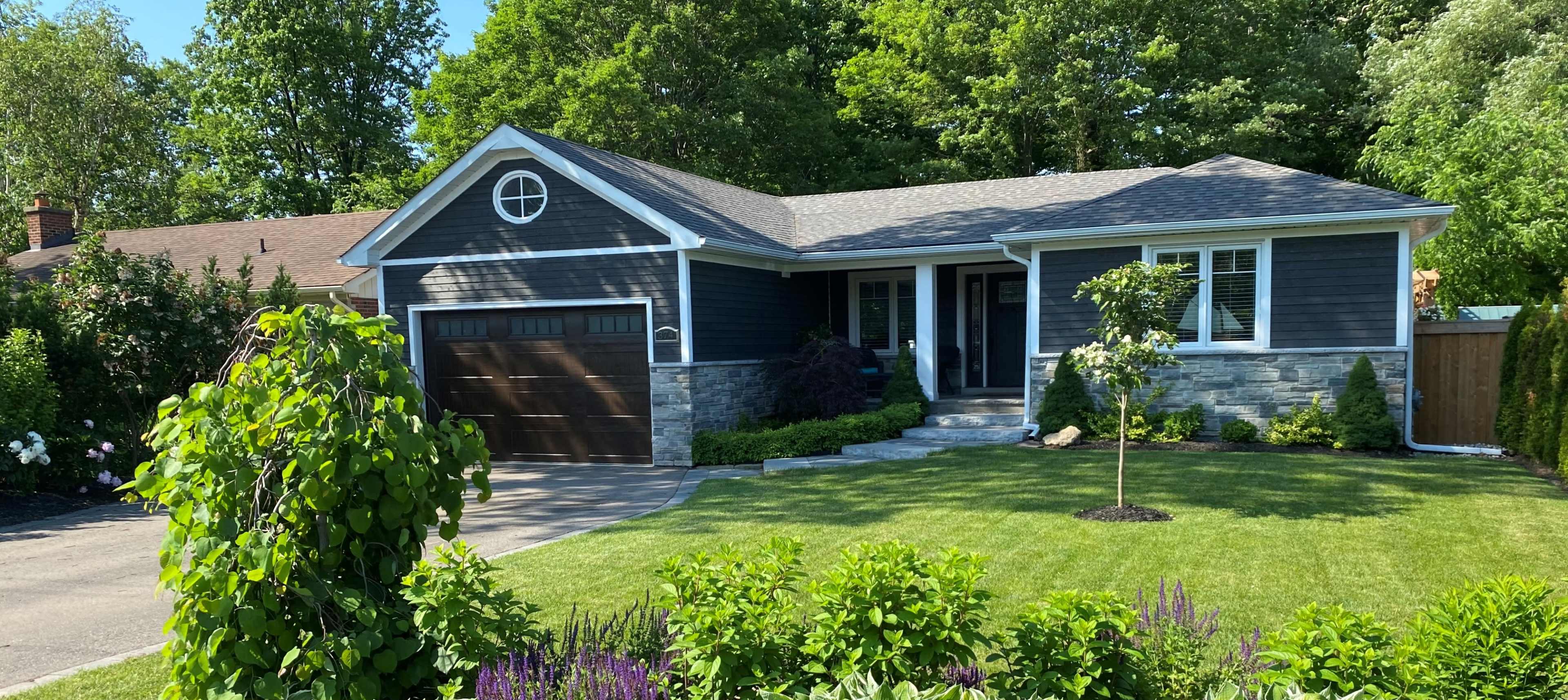 A single-story house with a dark exterior, a stone veneer, and a two-car garage is surrounded by green lawns and landscaping.