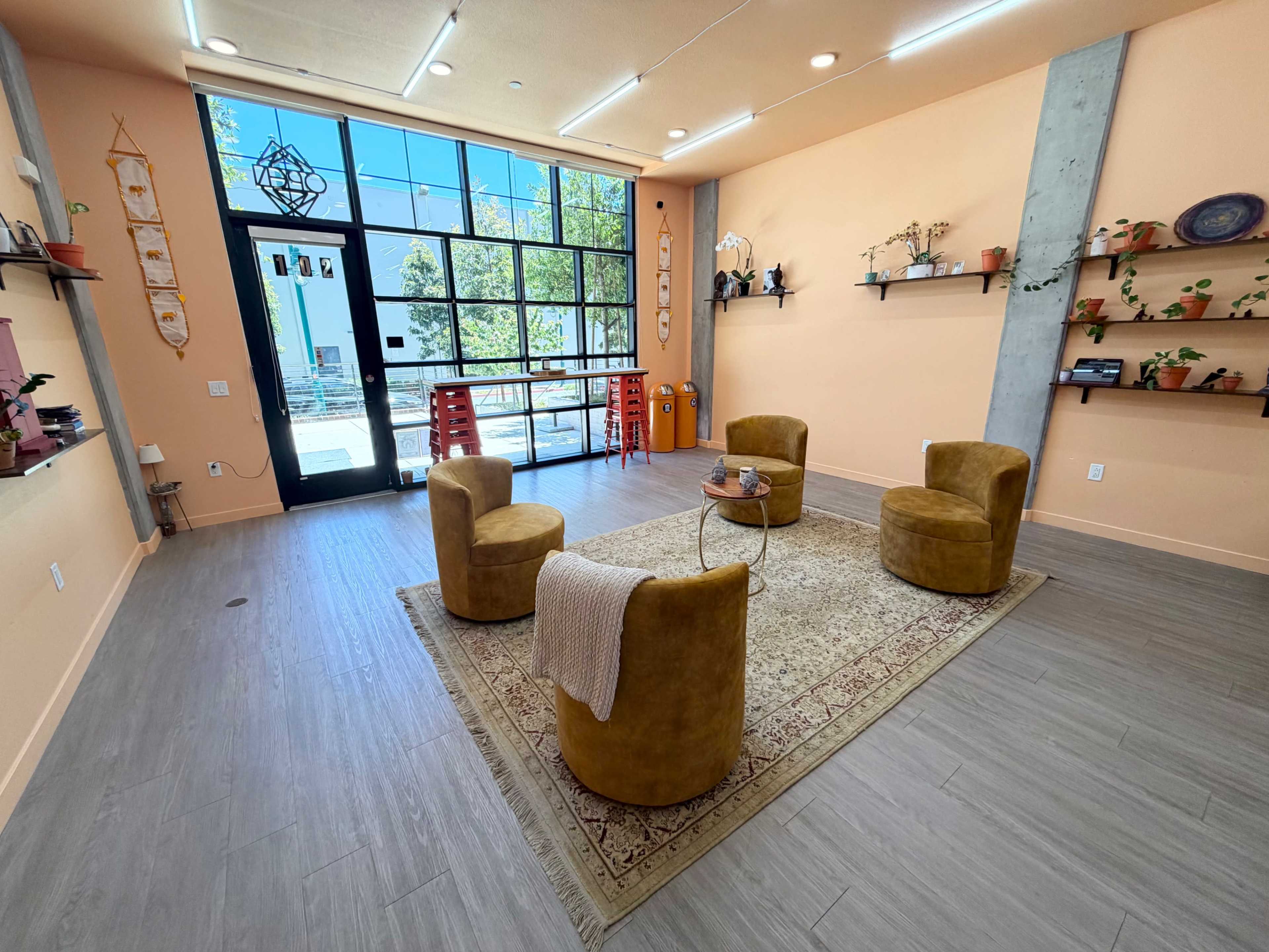 The image shows a cozy room with three yellow chairs arranged around a small table on a decorative rug, against a backdrop of light orange walls, large windows, and minimalistic shelving.