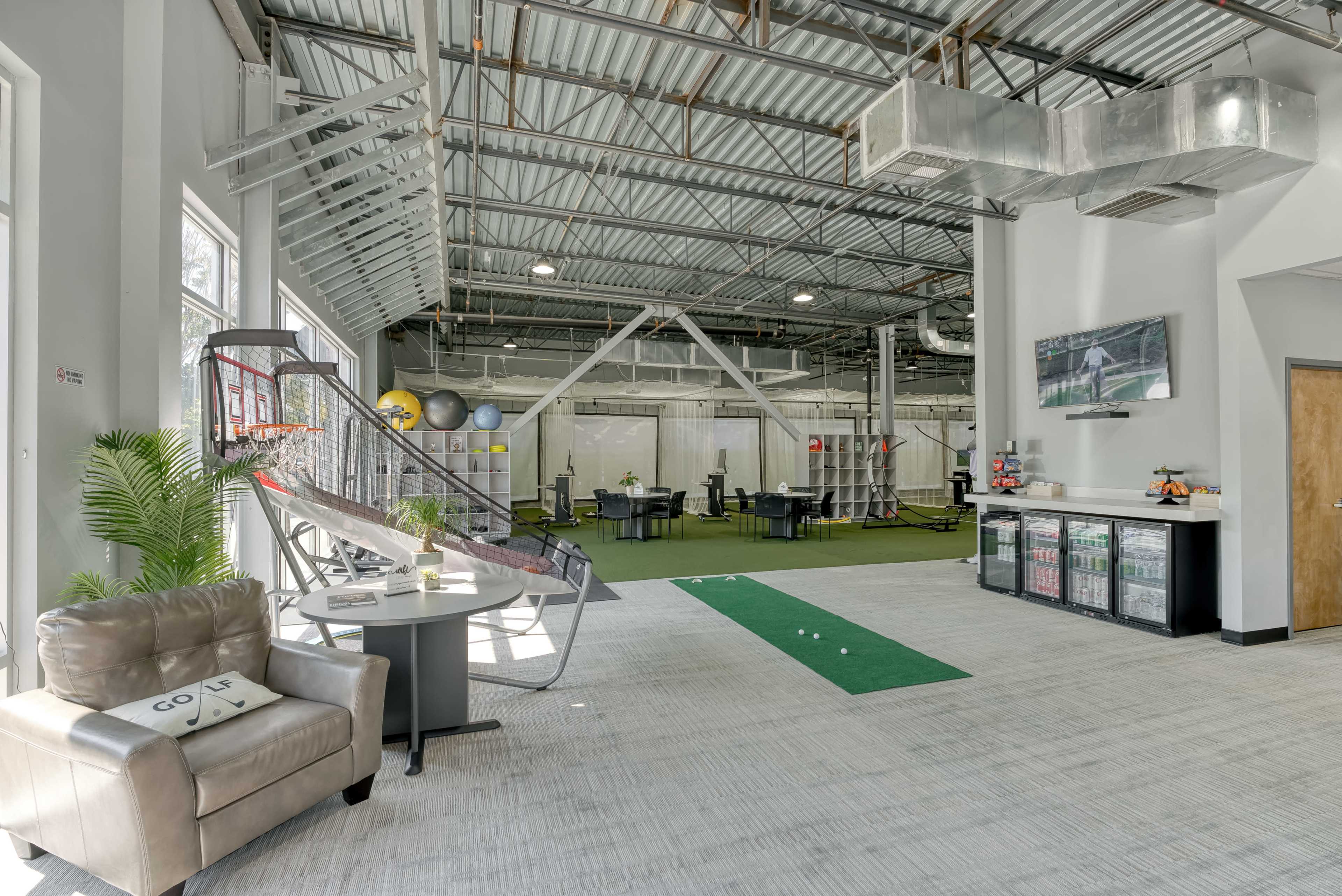A spacious indoor facility featuring areas for golf practice, seating, and a bar-like area with a selection of drinks.