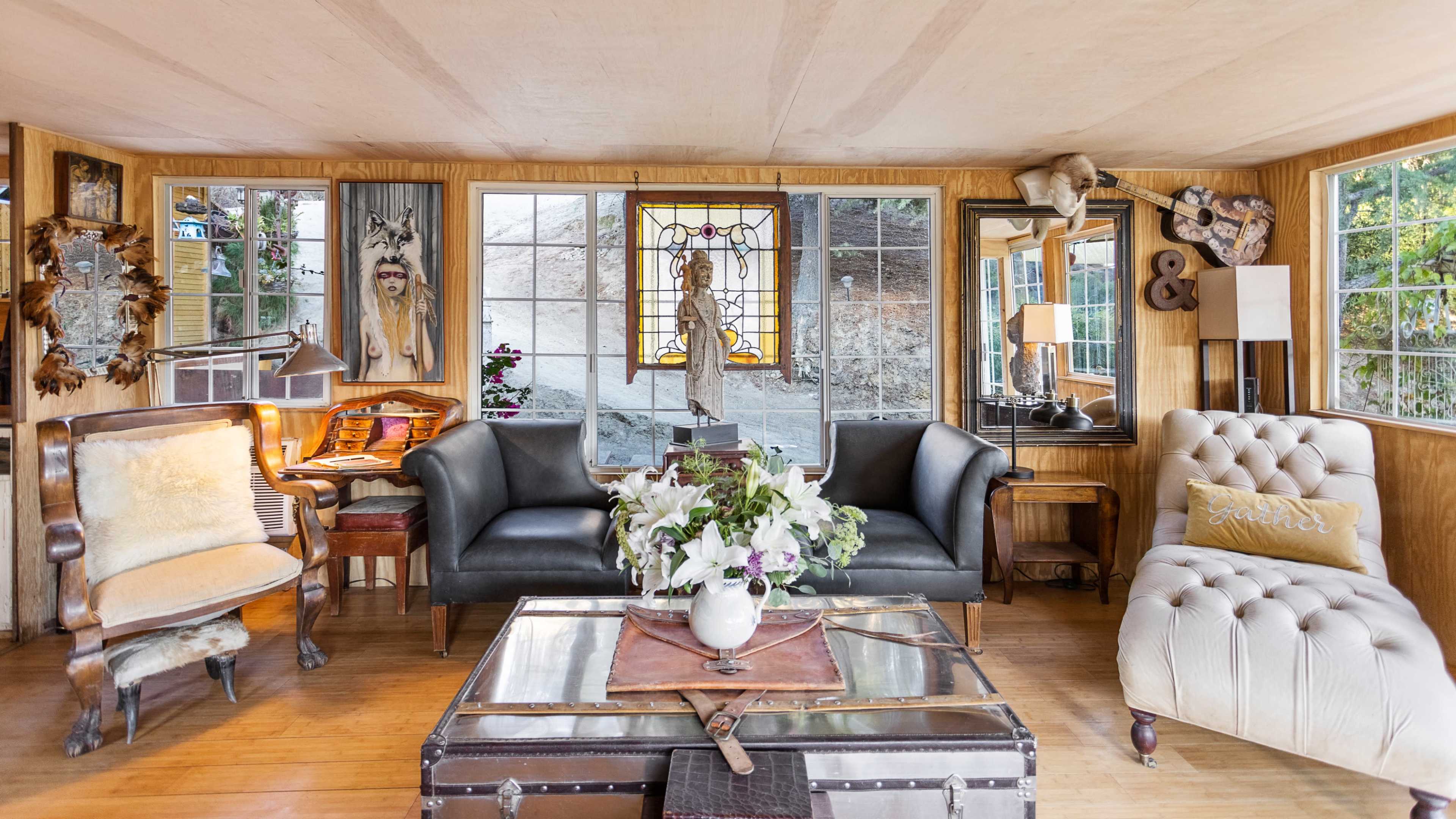 The image shows a cozy living room featuring two distinct sofas, a coffee table with a decorative centerpiece, large windows, and various artworks on the walls.