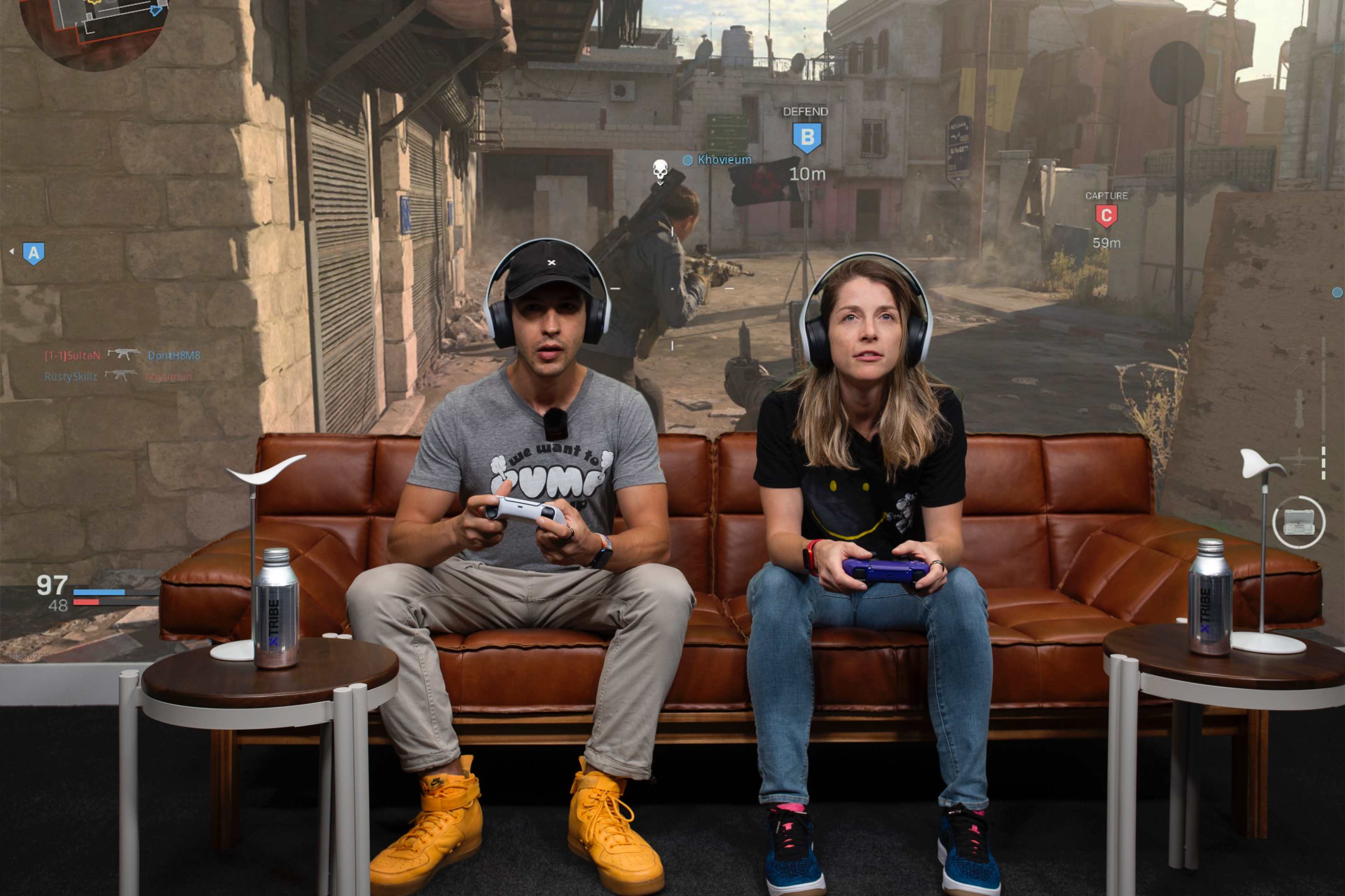 Two gamers sit on a brown couch with game controllers, focused on a video game displayed on a large screen in a gaming setup.