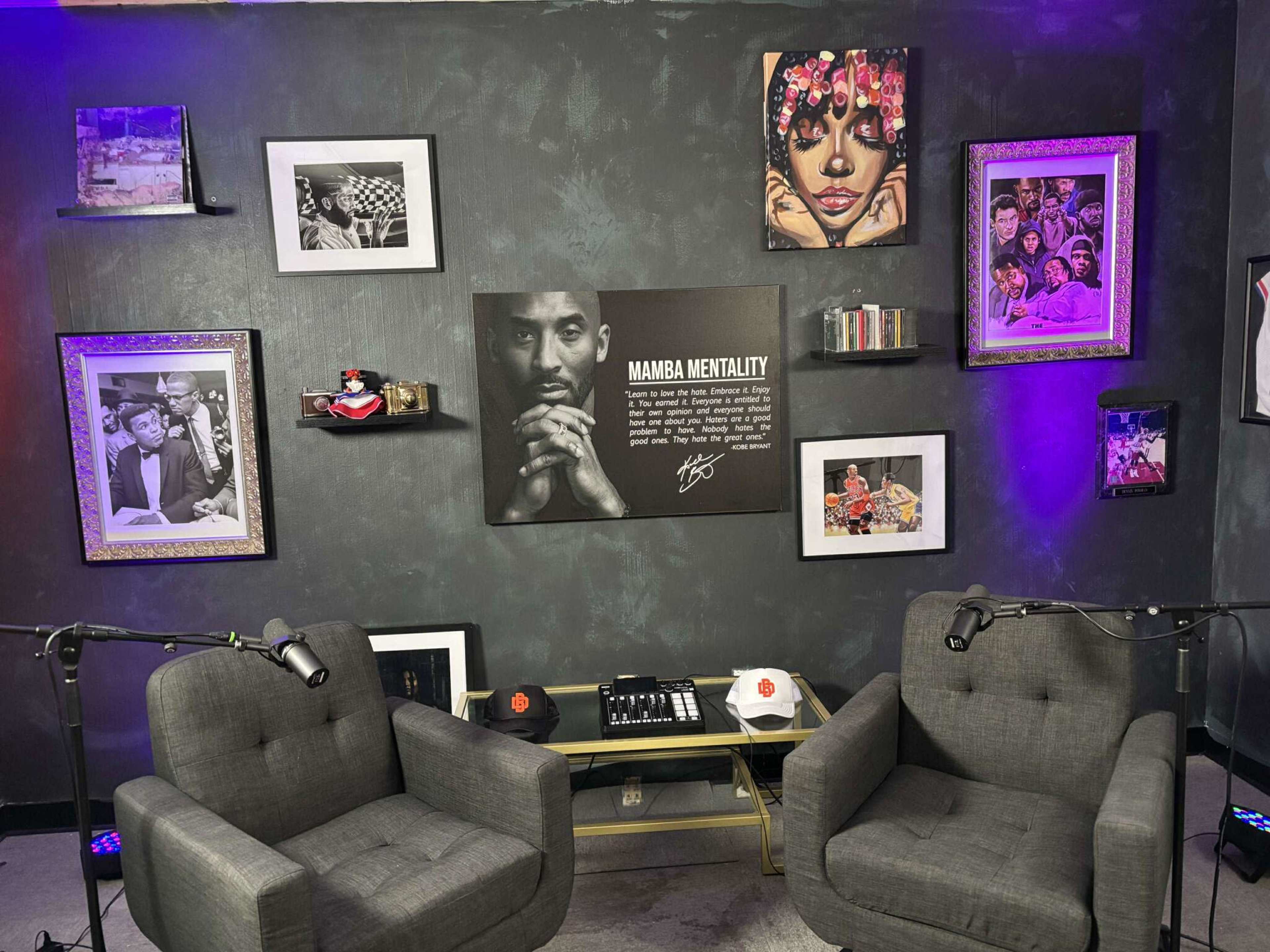 The image shows a podcast or interview setup with two gray chairs, a table with equipment, and several framed photographs and artwork on the walls, including a central piece featuring a well-known basketball player.
