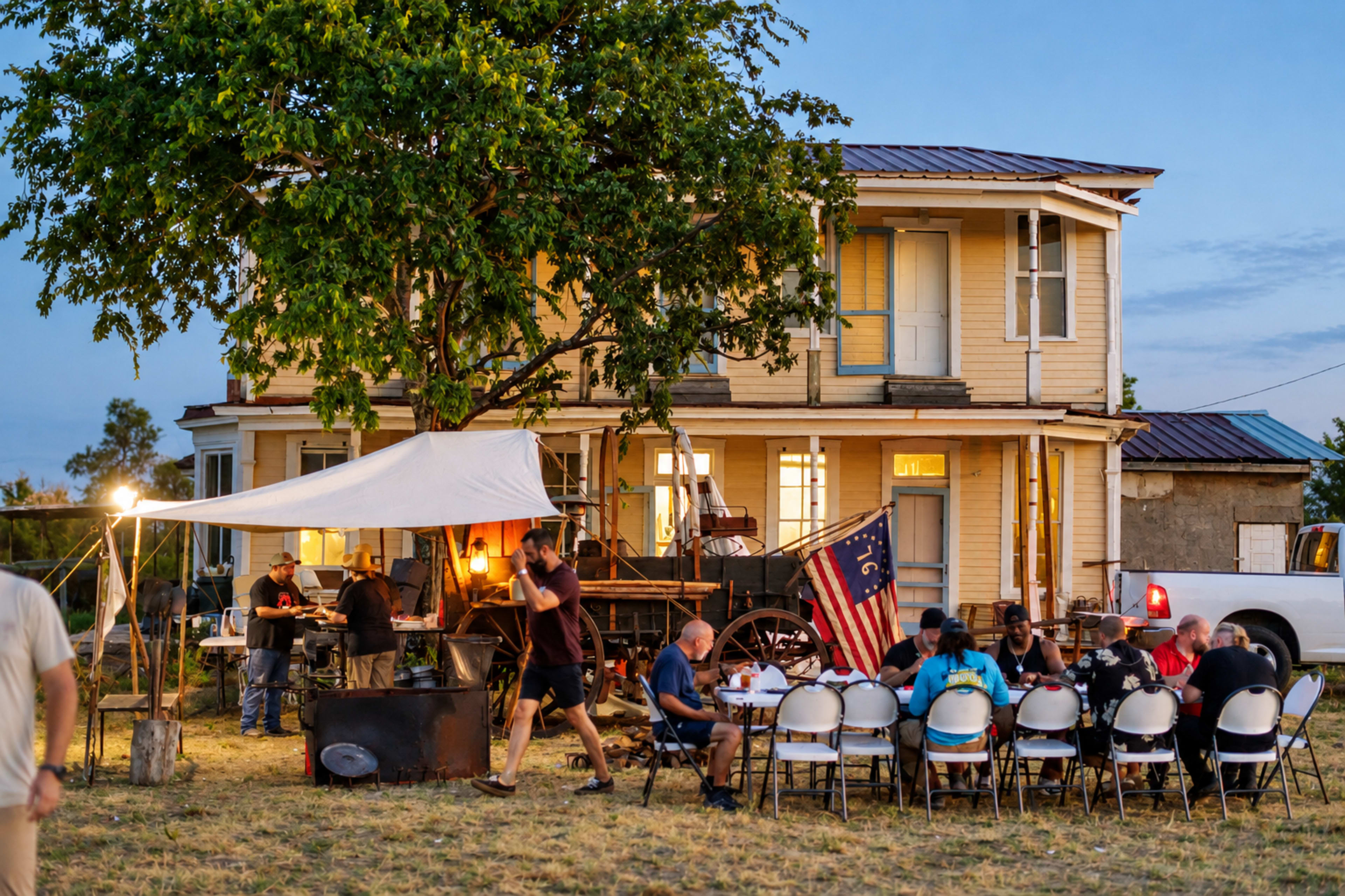 A group of people gather around tables for an outdoor meal near a two-story house with a large tree and an American flag.
