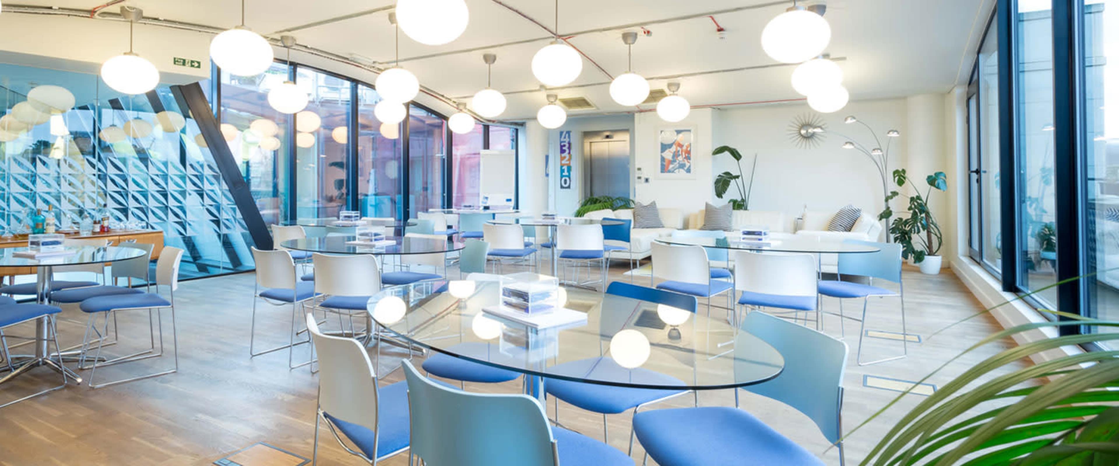 A modern office space with multiple glass tables and white chairs, featuring round pendant lights and large windows.