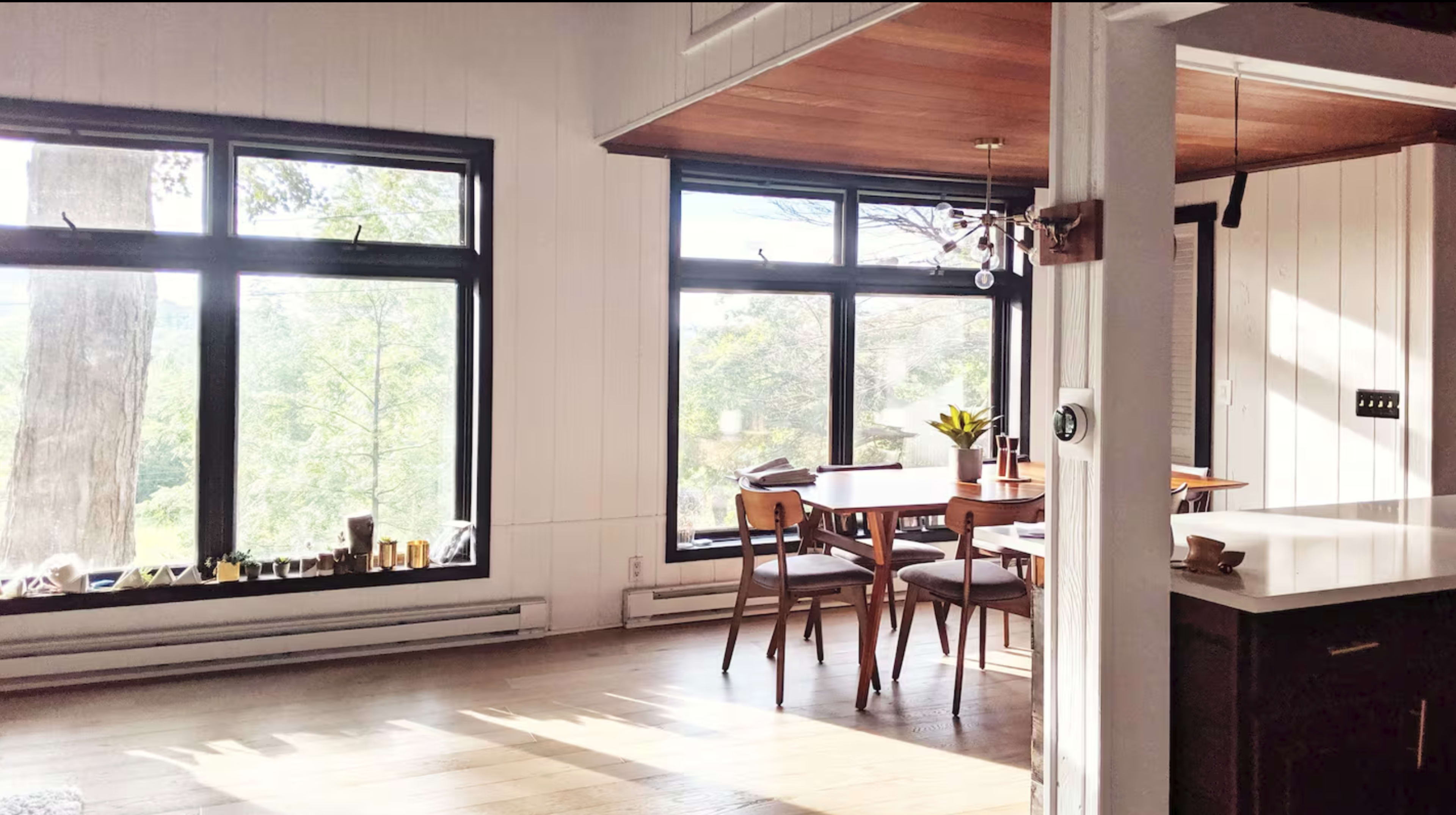 A bright, airy room features large windows overlooking greenery, a wooden dining table surrounded by chairs, and a modern kitchen area.