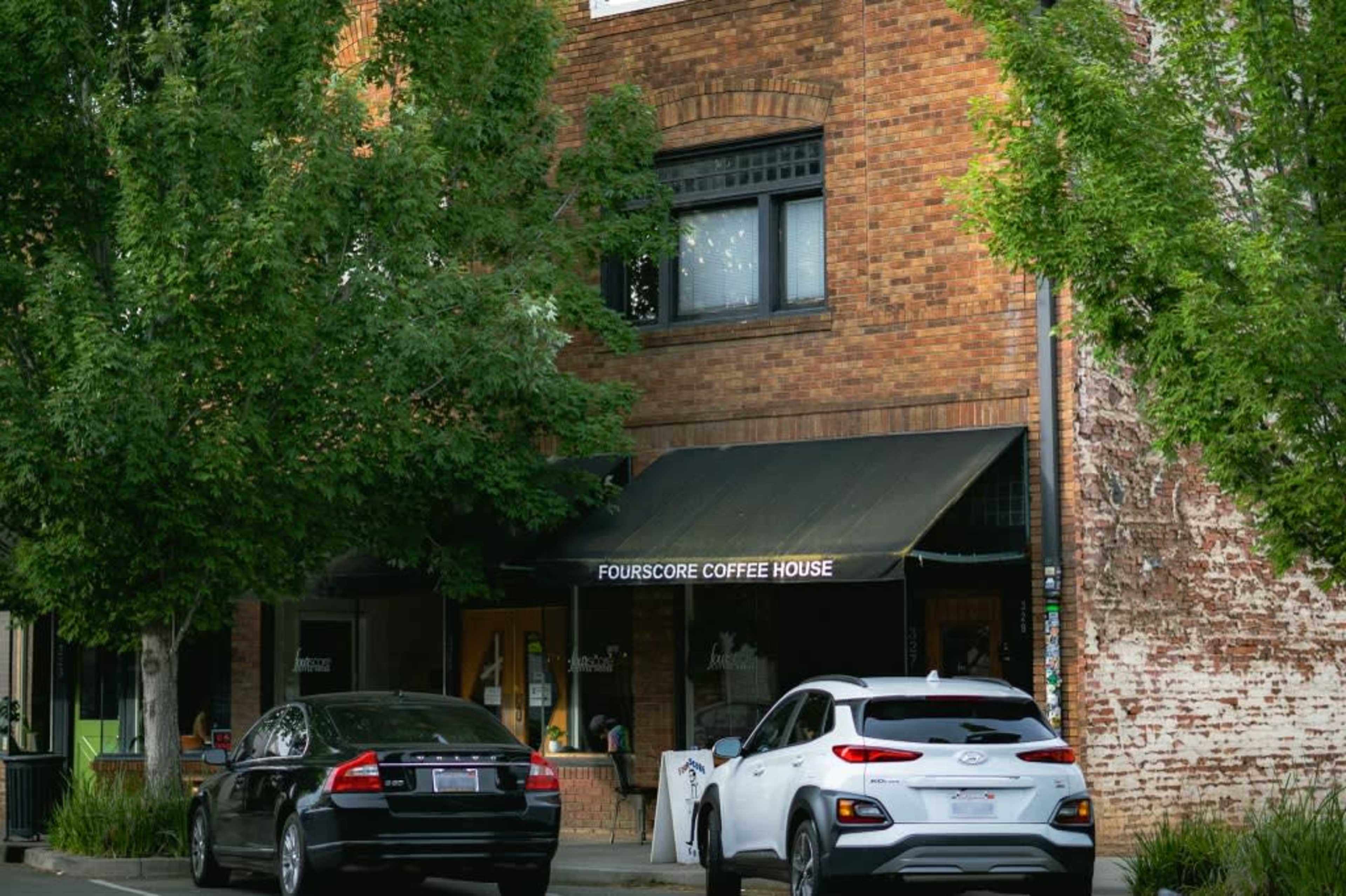 A brick building with a black awning labeled "FOURSCORE COFFEE HOUSE," flanked by two parked cars and surrounded by trees.