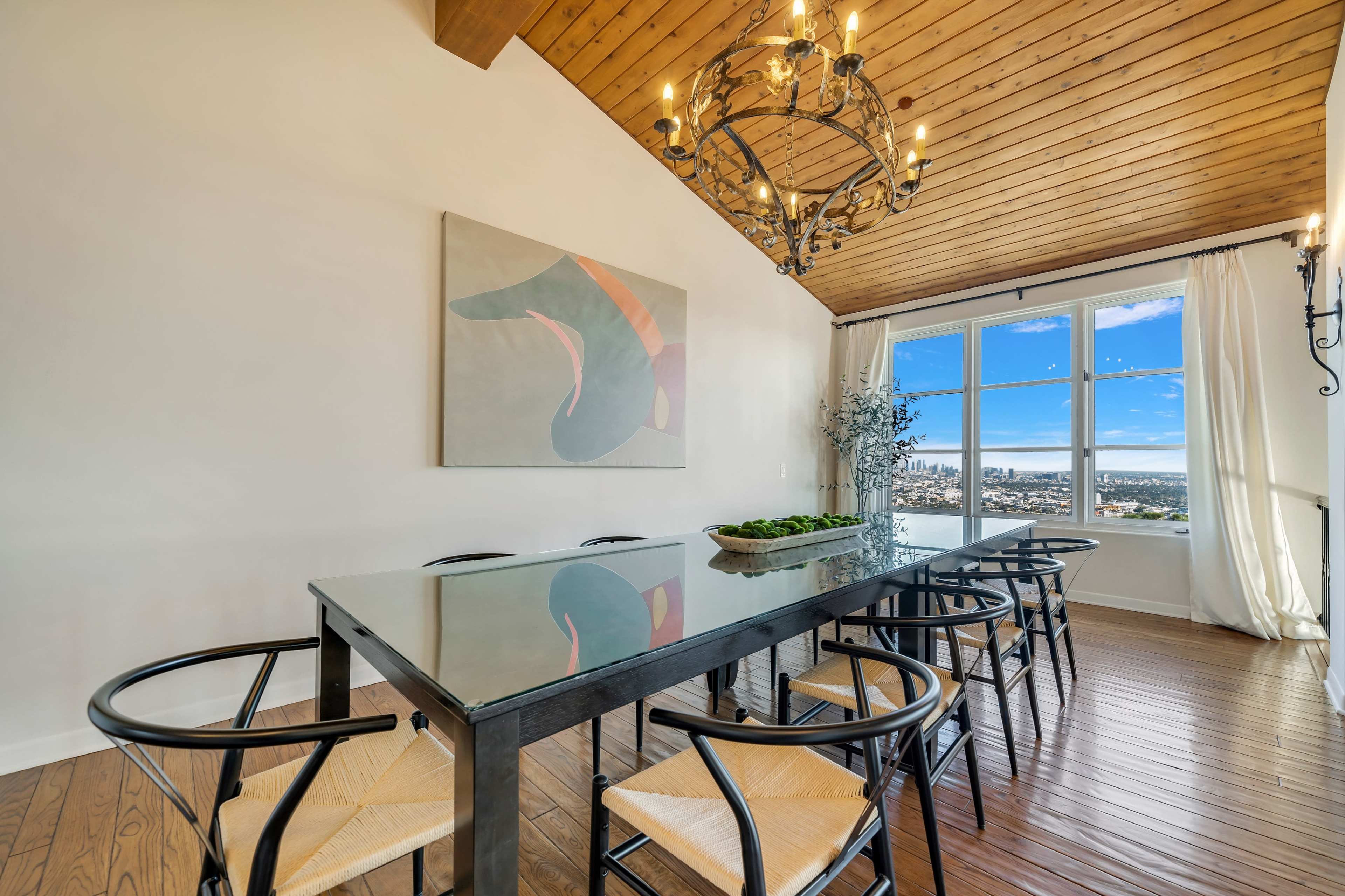 A dining room features a glass table surrounded by wooden chairs, with a chandelier above and large windows displaying a city view.