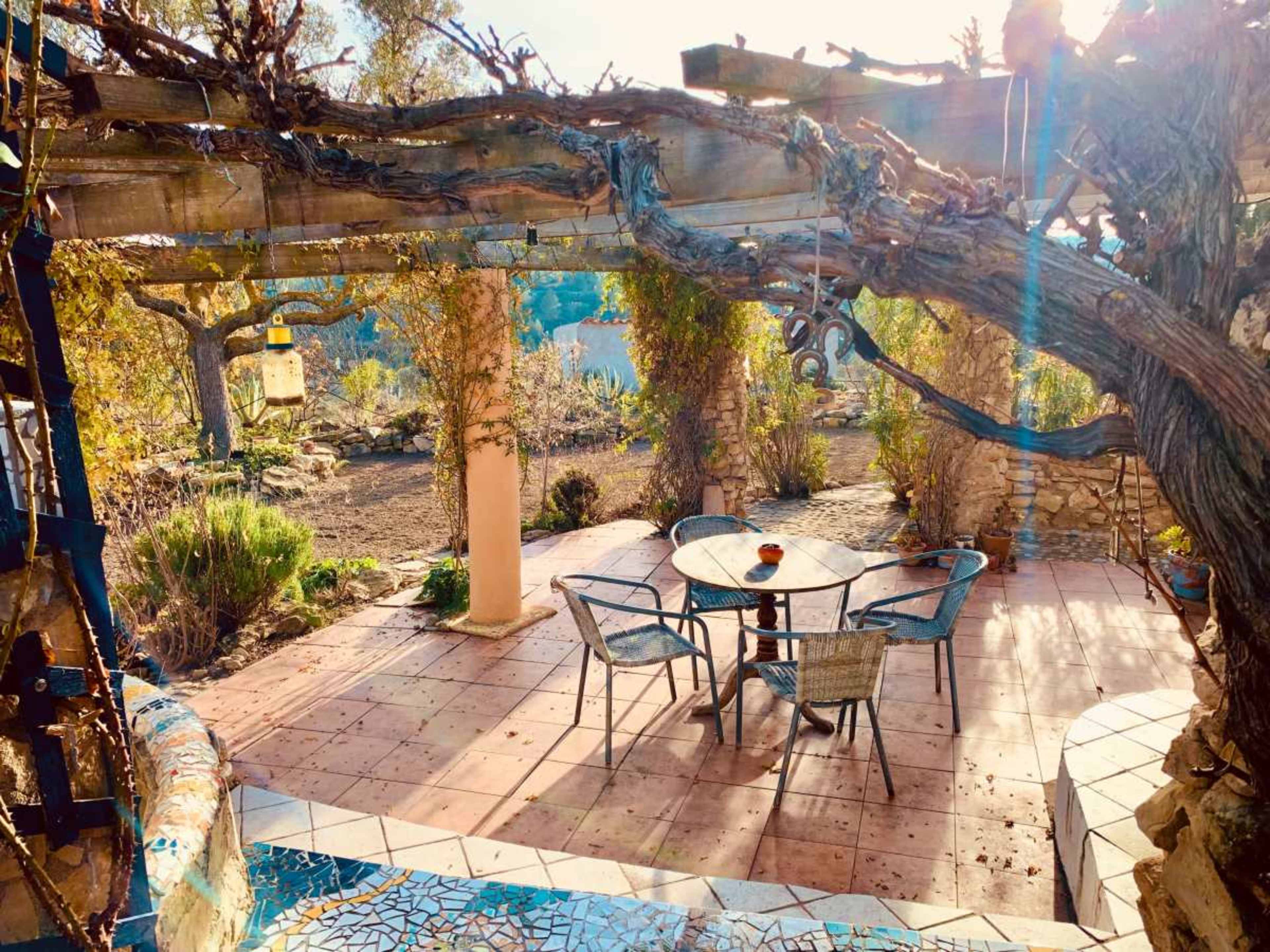 A patio dining area is surrounded by lush greenery, featuring a round table and chairs under a wooden pergola.