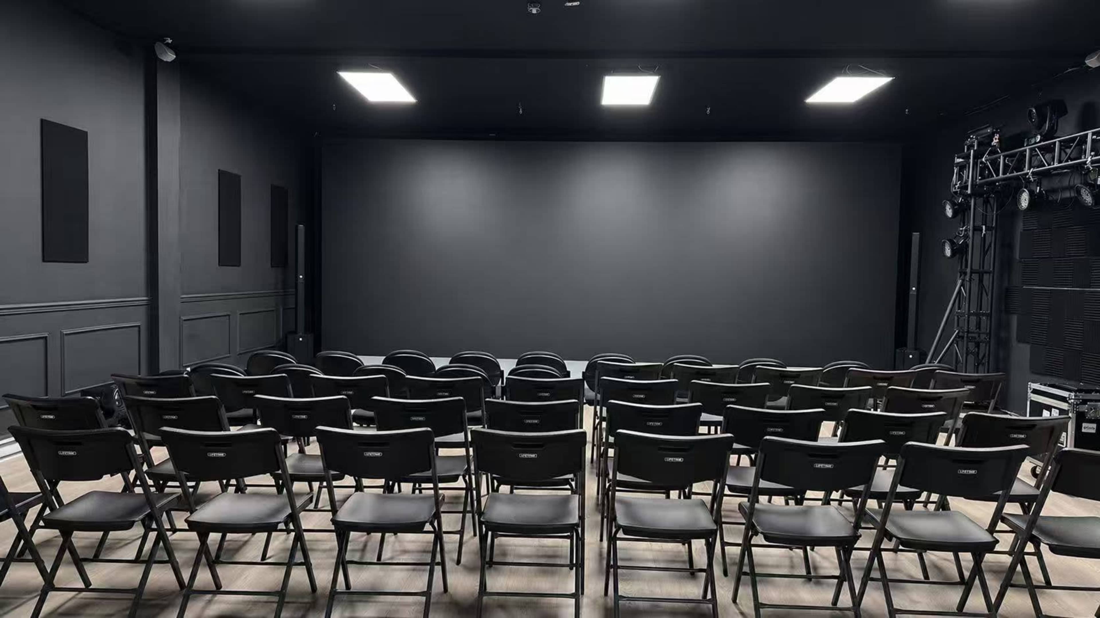 A row of black folding chairs is arranged in front of a large blank screen in a dimly lit auditorium.