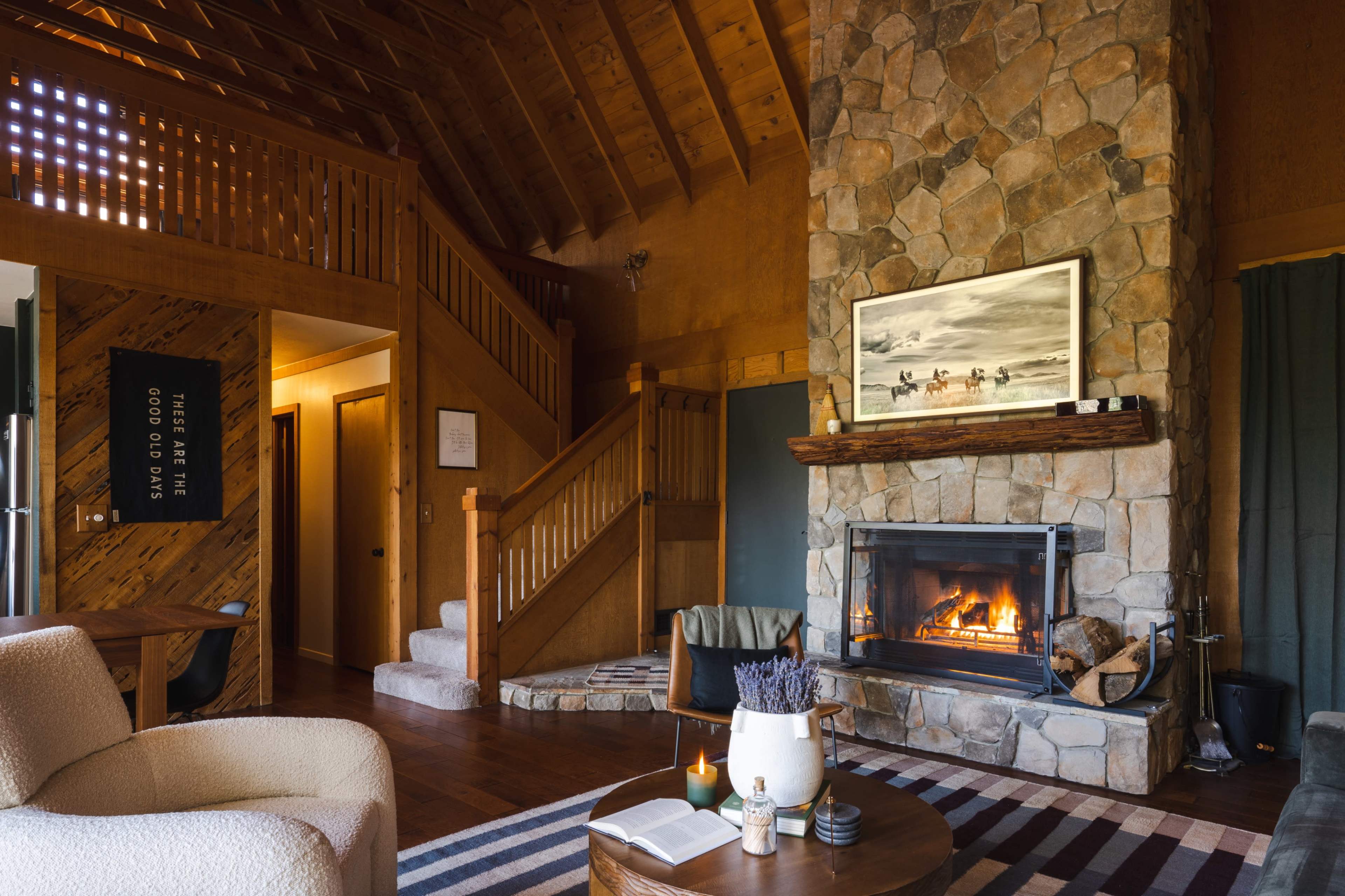 A cozy interior living space featuring a stone fireplace, wooden beams, a staircase, and a comfortable seating area.