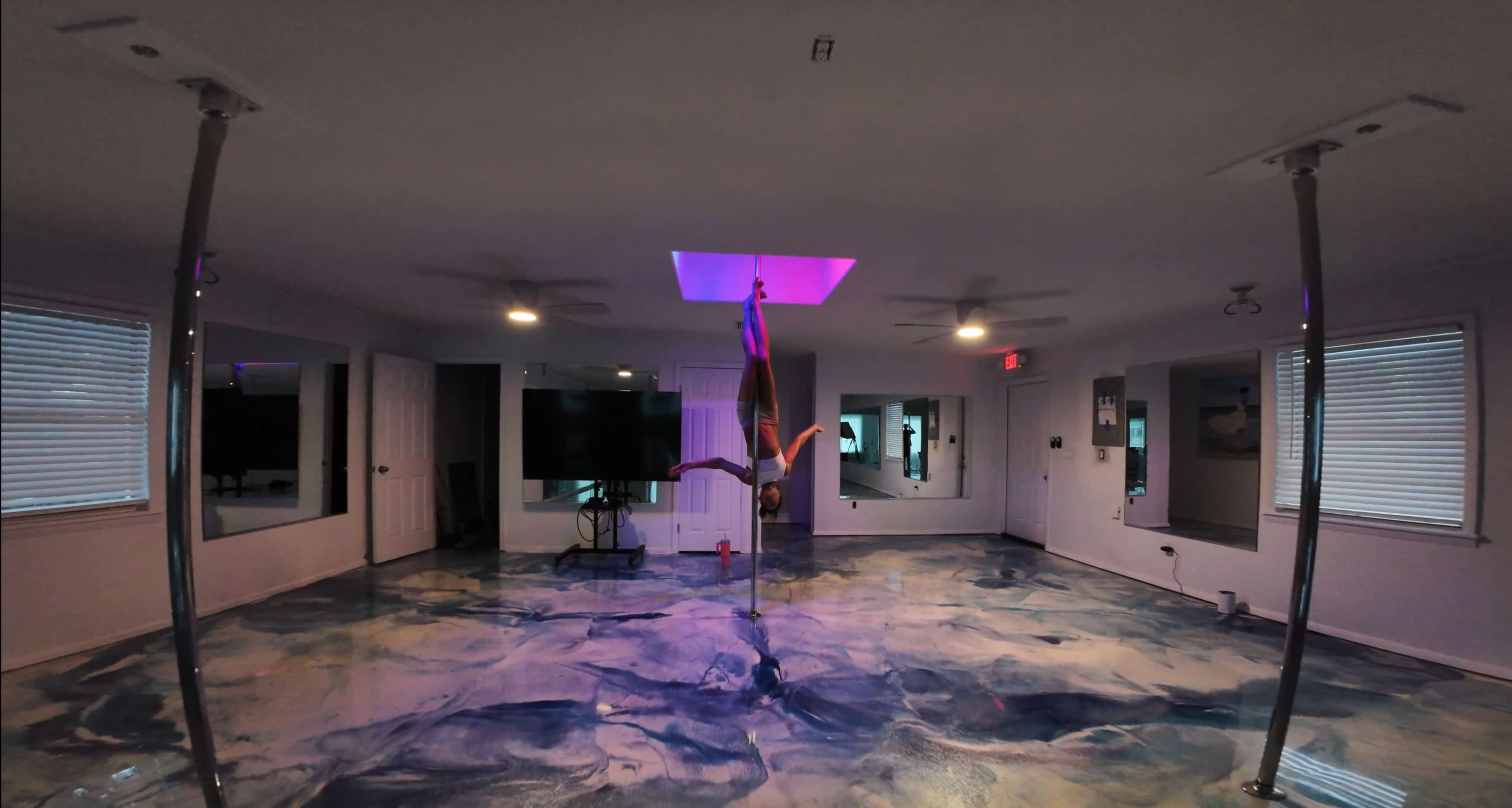 A person performs on a pole in a brightly lit room with an artistic floor design and multiple windows.