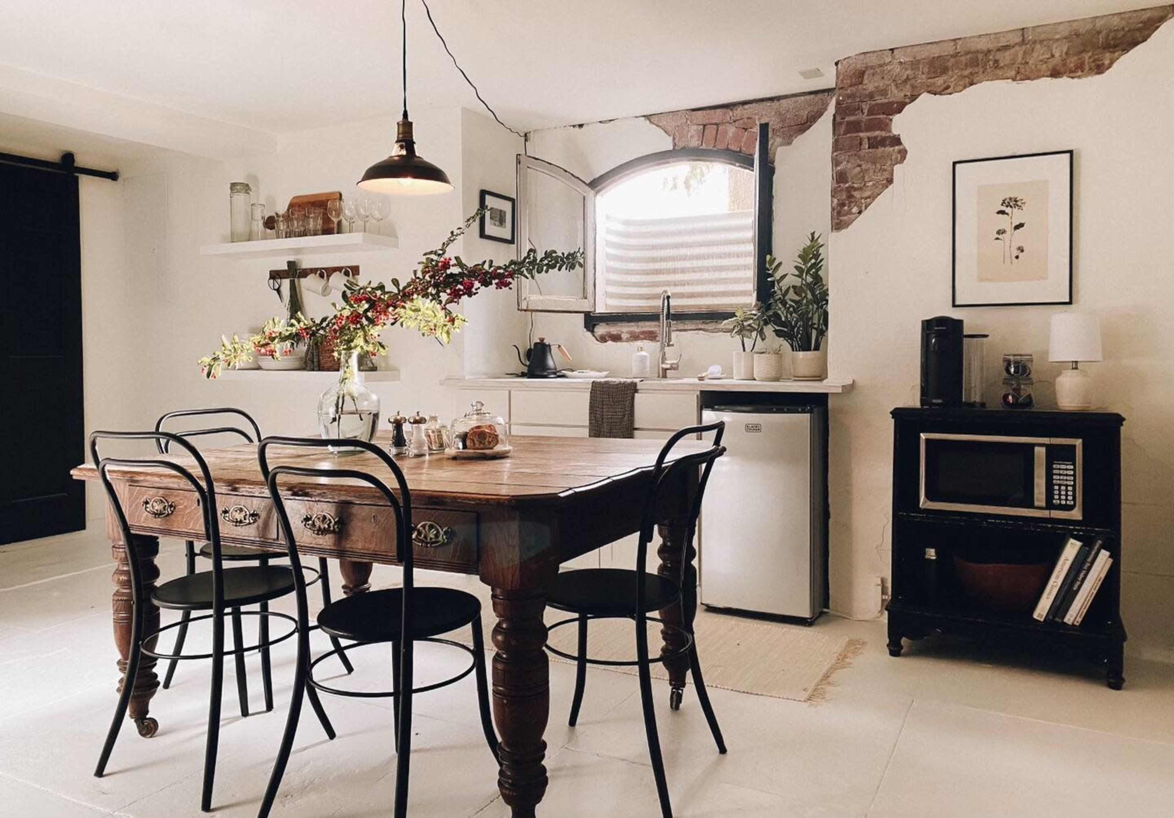 A rustic kitchen features a wooden table surrounded by black chairs, with a vase of flowers and various kitchen appliances visible in the background.