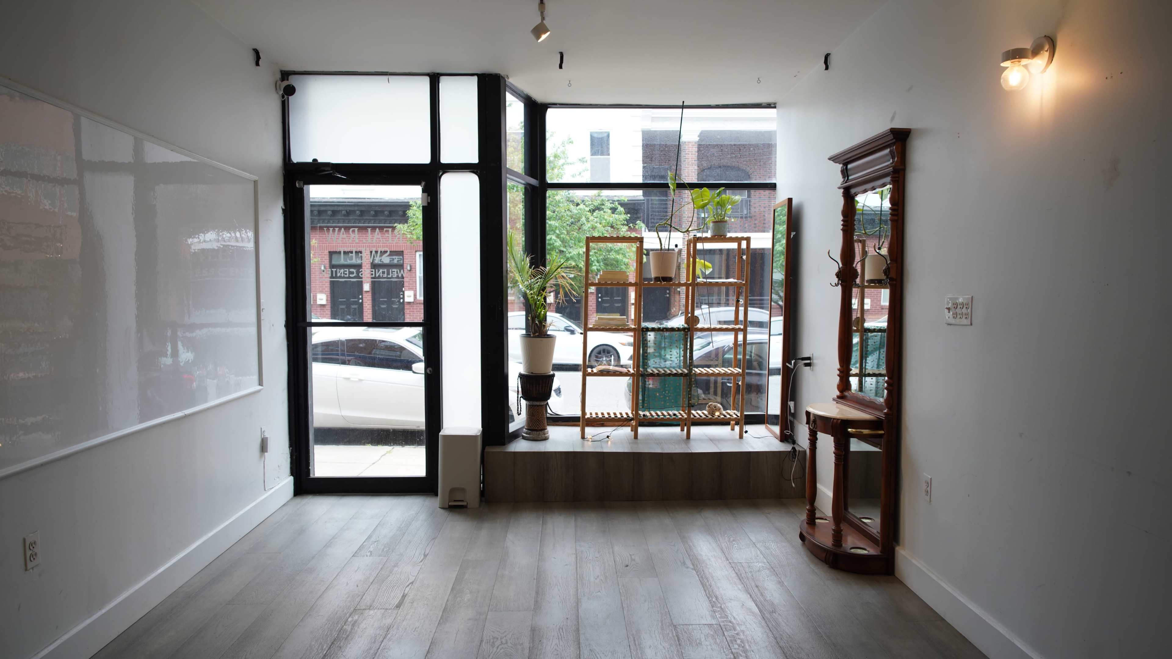 The image shows an empty commercial space with large windows facing a street, featuring a white wall, wooden flooring, and minimal furnishings including a display rack and a small table.
