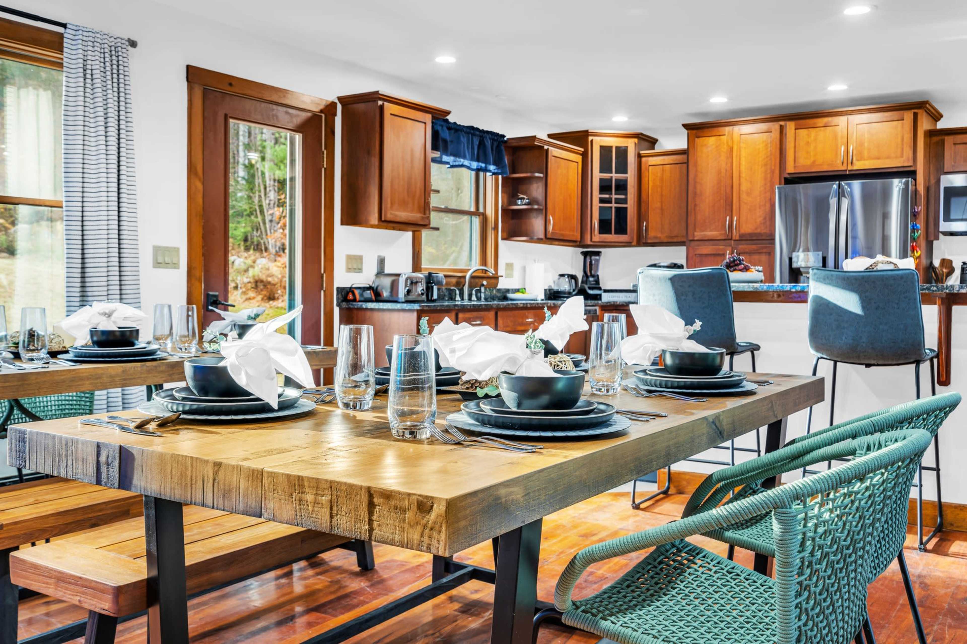 A modern dining area features a wooden table set with dark plates and white napkins, surrounded by turquoise chairs, with an open kitchen in the background.
